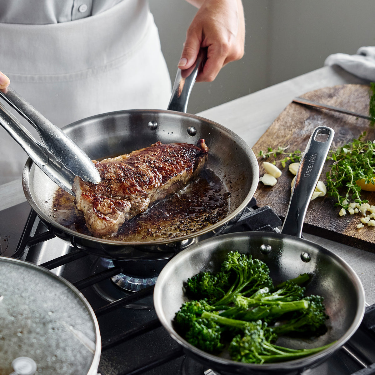 Chatham Pro 12-piece stainless steel skillet searing a steak with tongs, with broccoli in a nearby pan.