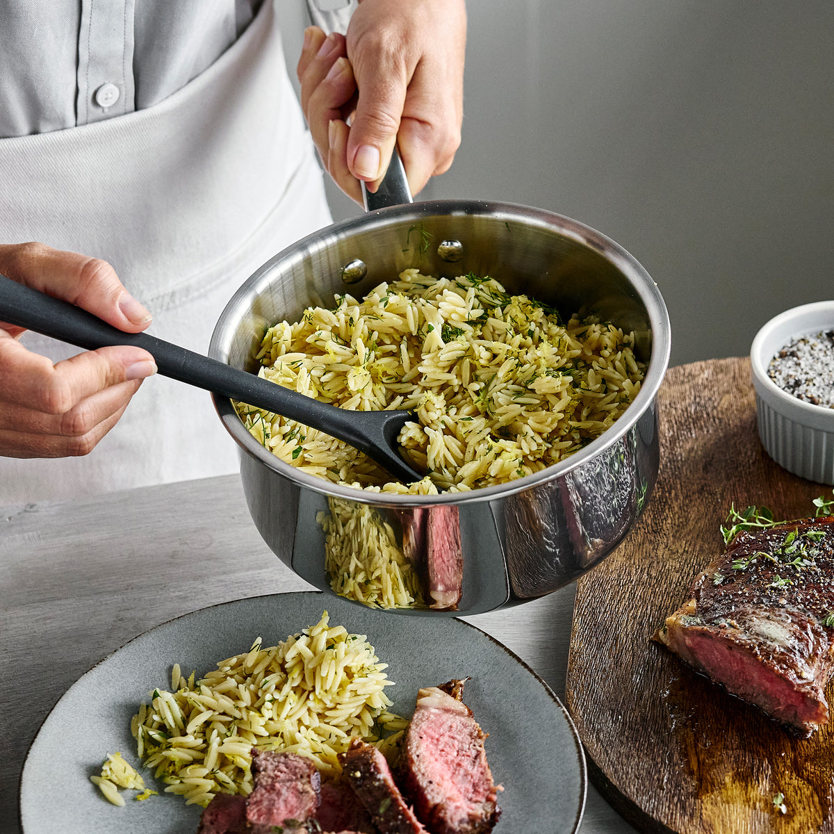 Hands holding a Chatham Pro stainless steel saucepan, serving orzo and sliced steak onto a plate.