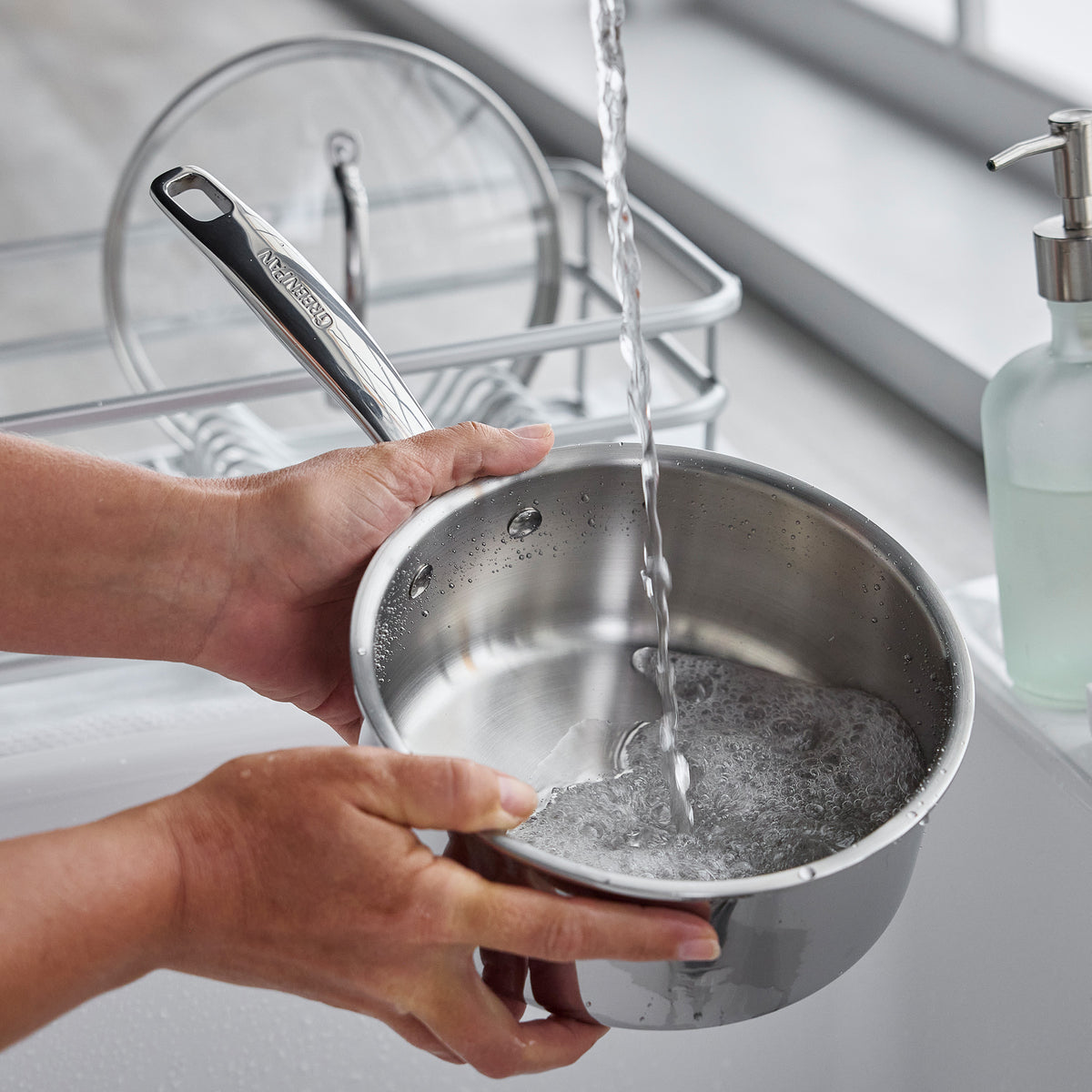 Hands washing a Chatham Pro stainless steel saucepan from the 12-piece cookware set under a kitchen faucet.