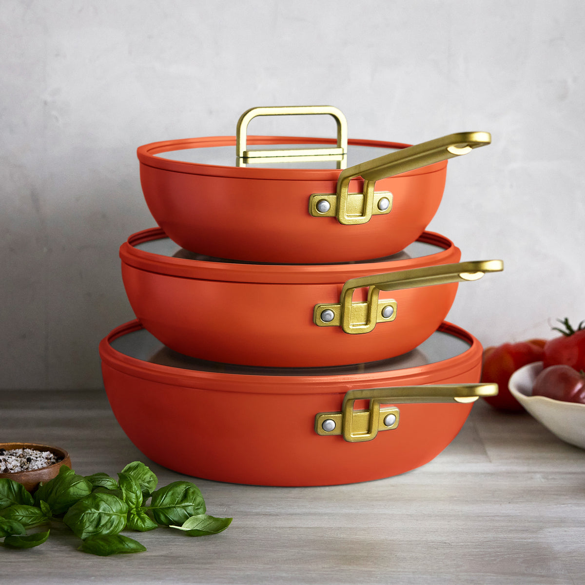 Three 3.25-Qt Stanley Tucci Piccolo pans in Amalfi Sunset with gold handles and glass lids, stacked on a countertop.