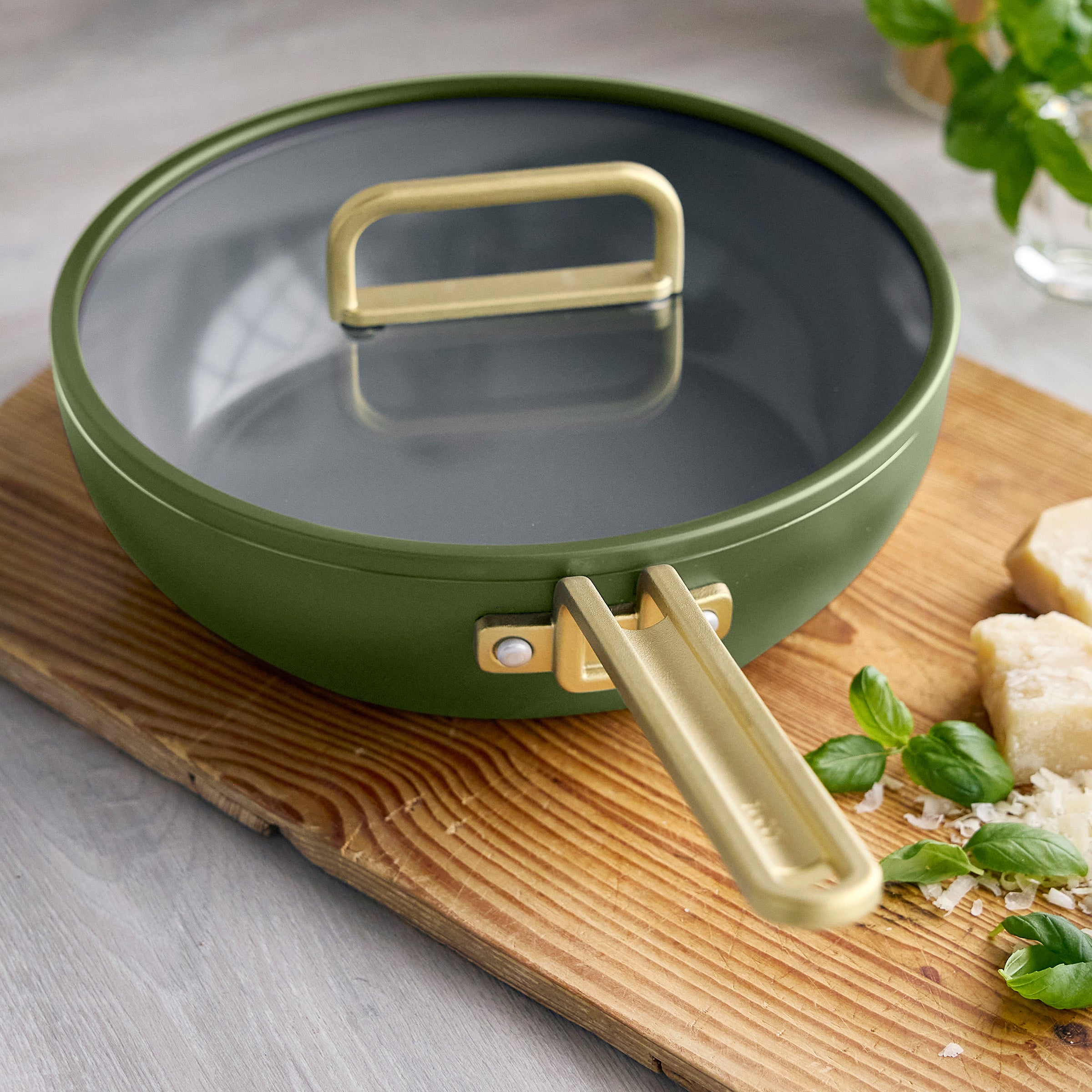 Ceramic nonstick piccolo pan in olive green on a wooden board, with fresh basil and cheese beside it.