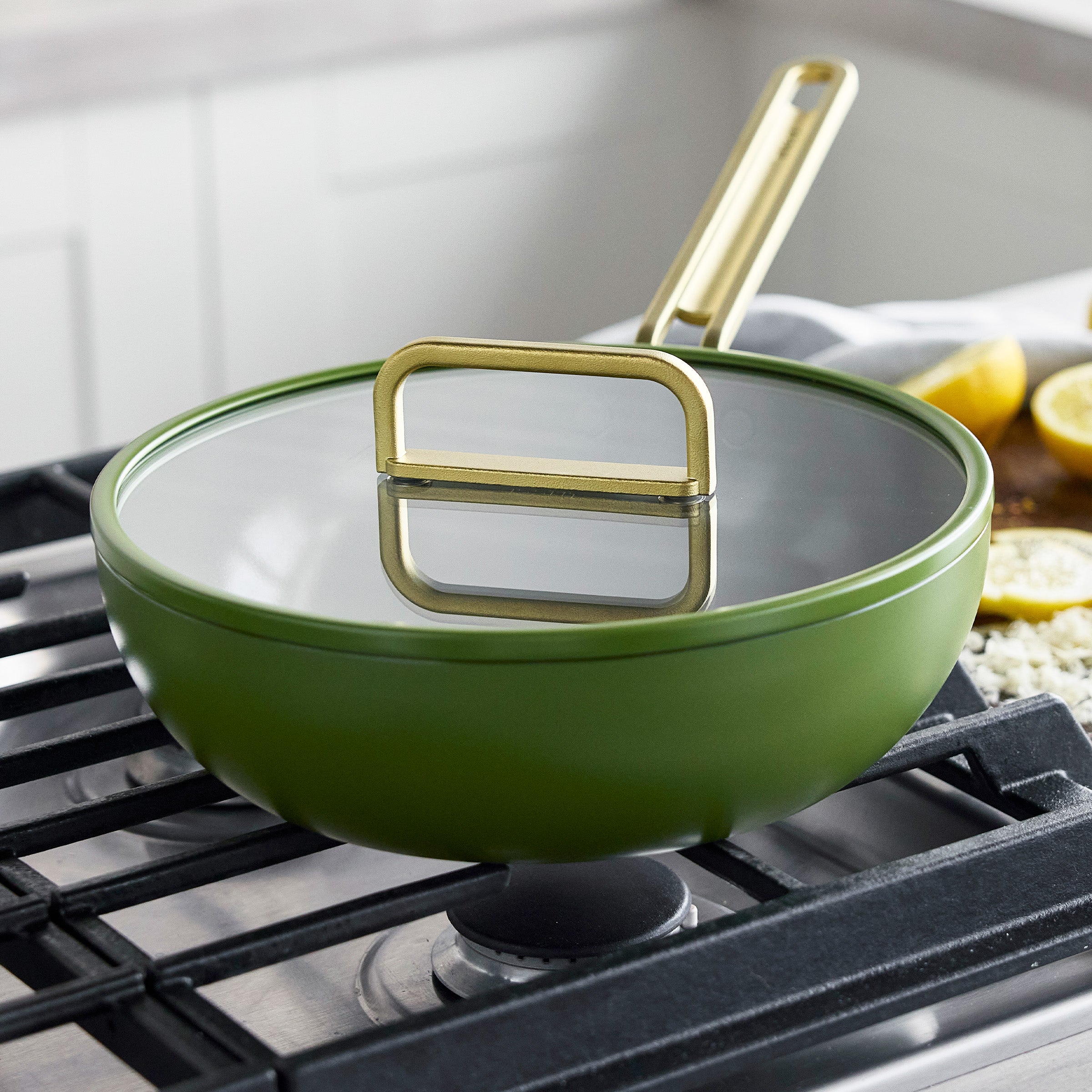 Ceramic nonstick piccolo pan in olive green with gold handle and lid on a stove, lemons and garlic beside it.