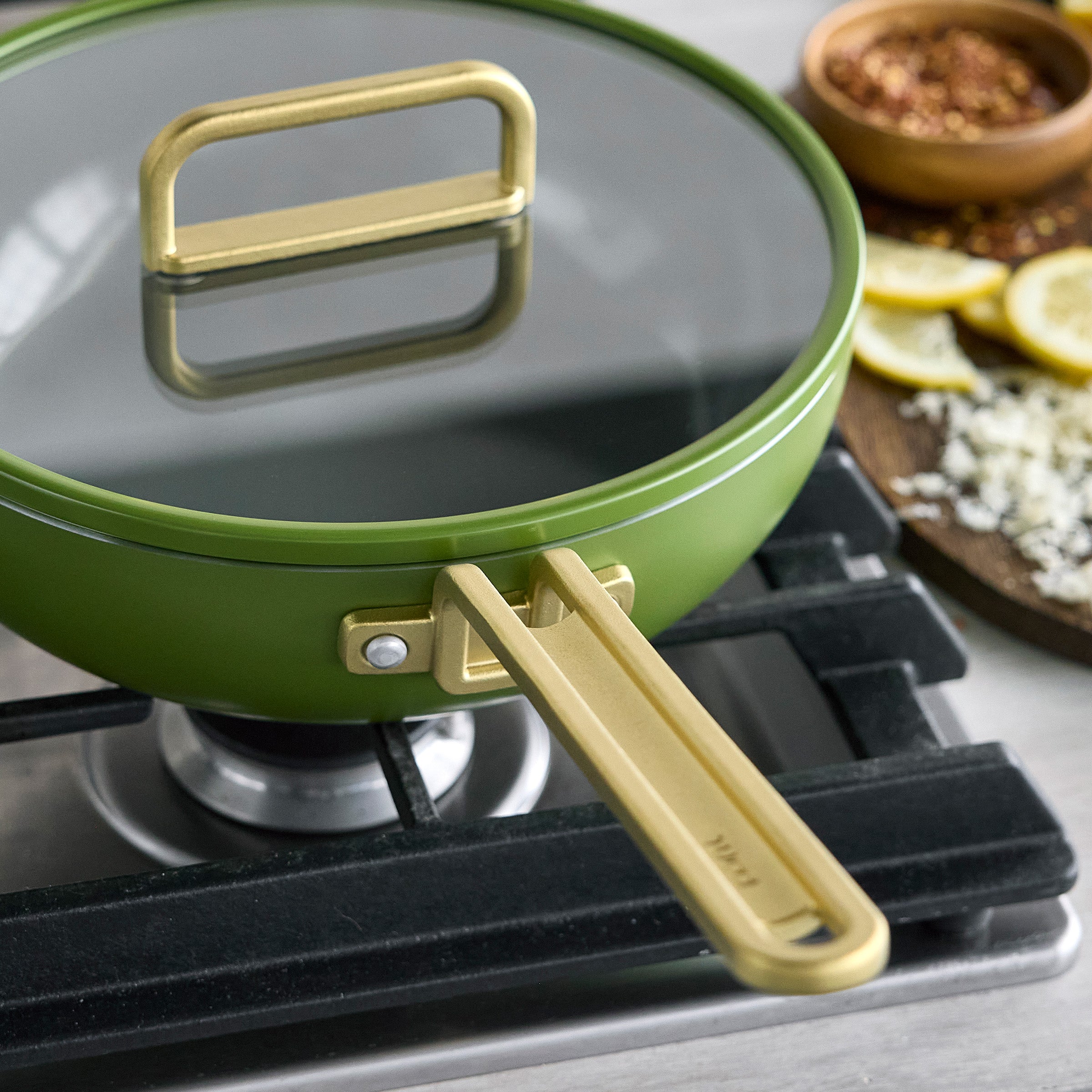 Green ceramic nonstick pan on a stove with lemons and spices on a wooden board nearby.