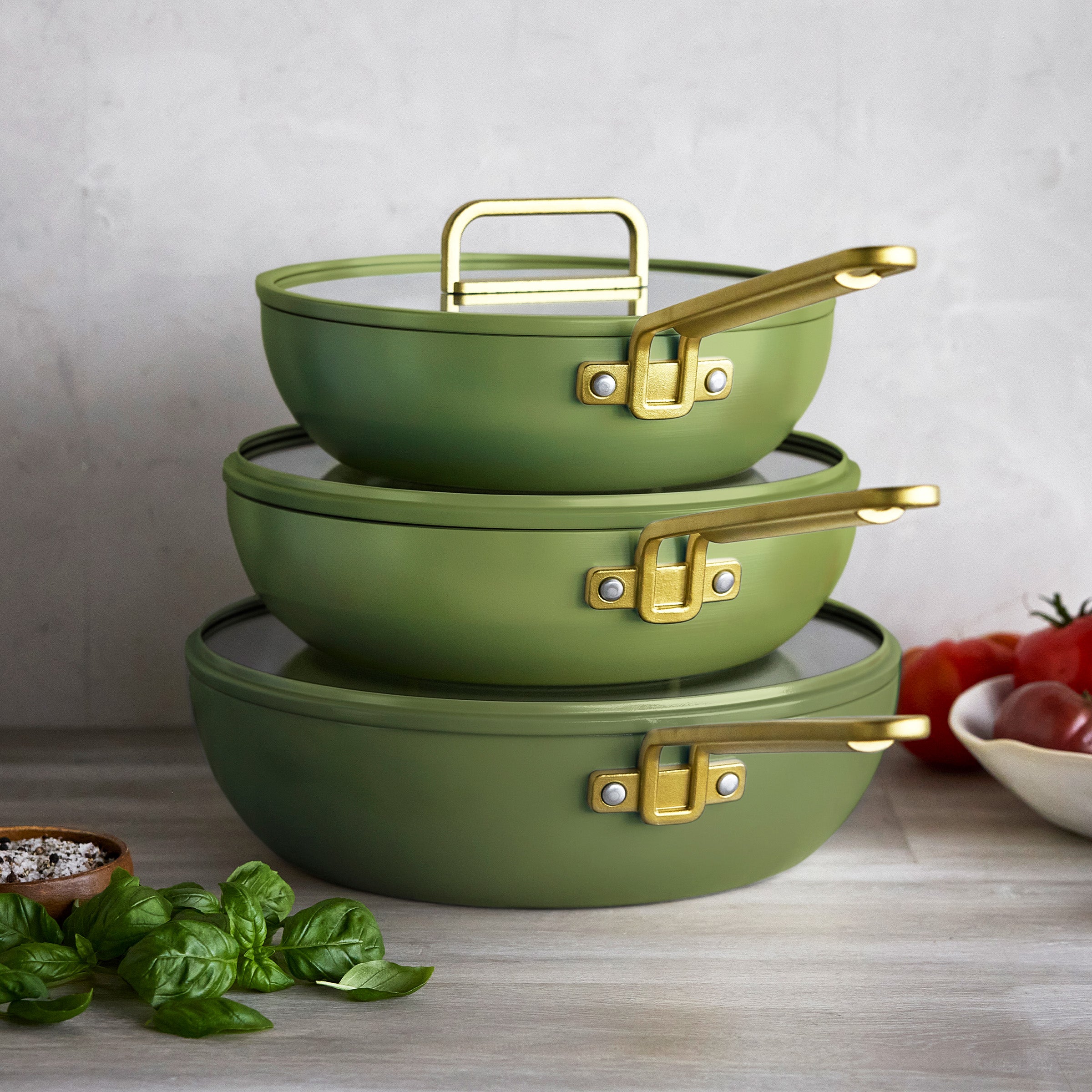 Set of three olive green Stanley Tucci Piccolo pans stacked on a counter with basil, tomatoes, and a small bowl nearby.