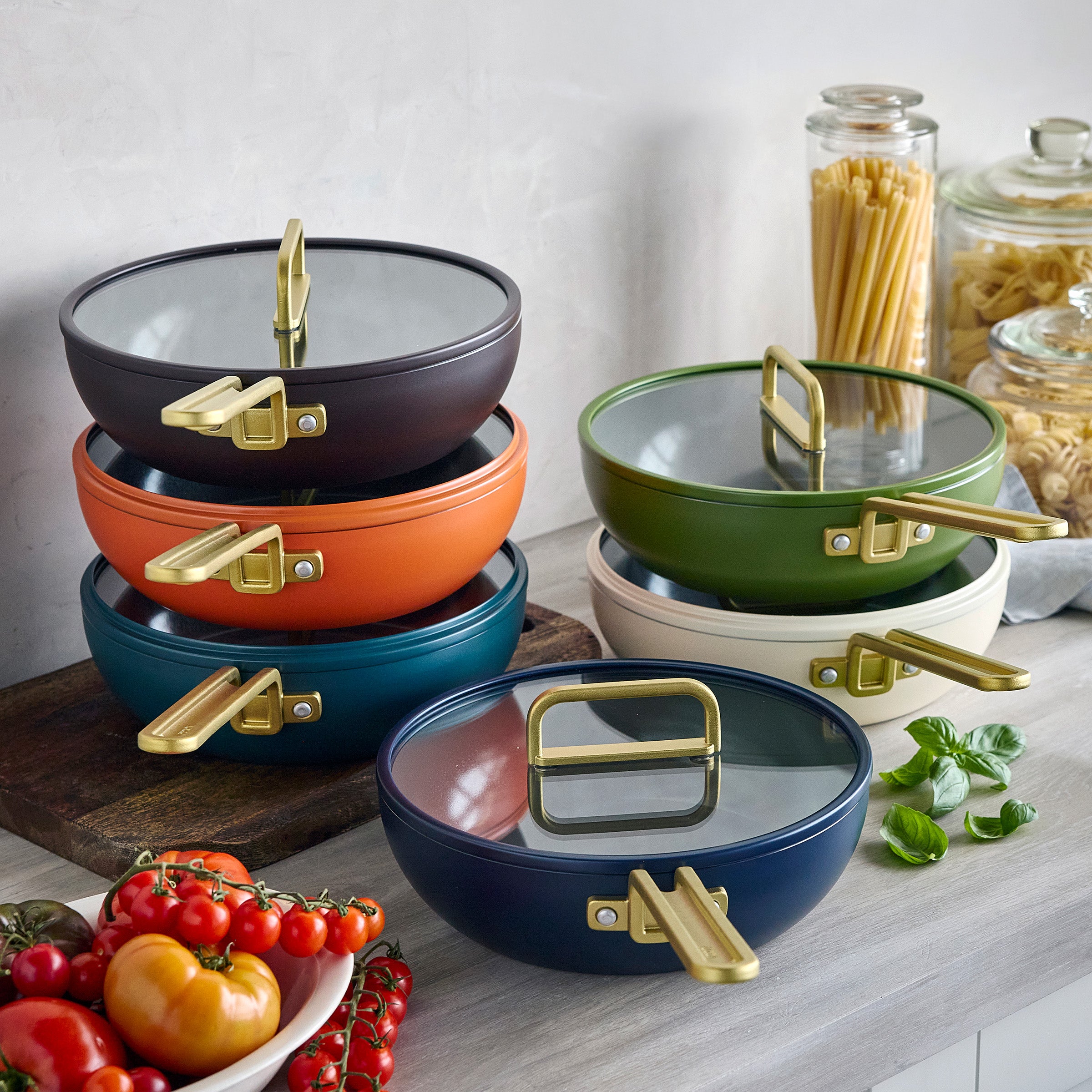 Six green Piccolo pans stacked on a kitchen counter near tomatoes, basil, and jars of pasta.