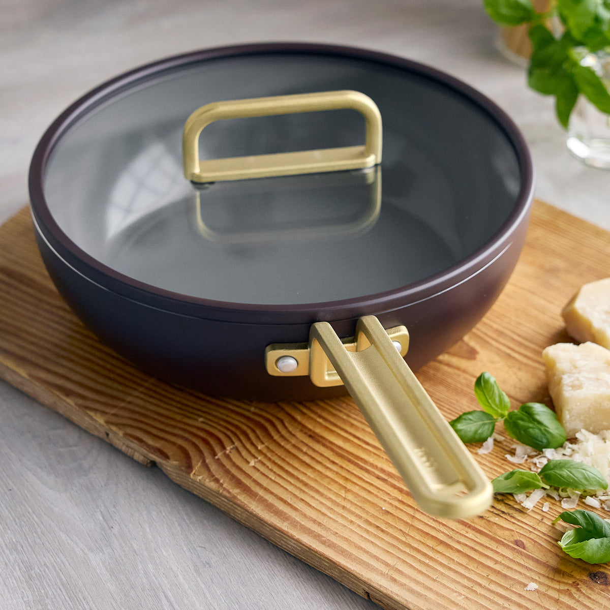 Espresso 3.25-quart ceramic nonstick Piccolo pan with glass lid and gold-tone handle on a wooden cutting board.