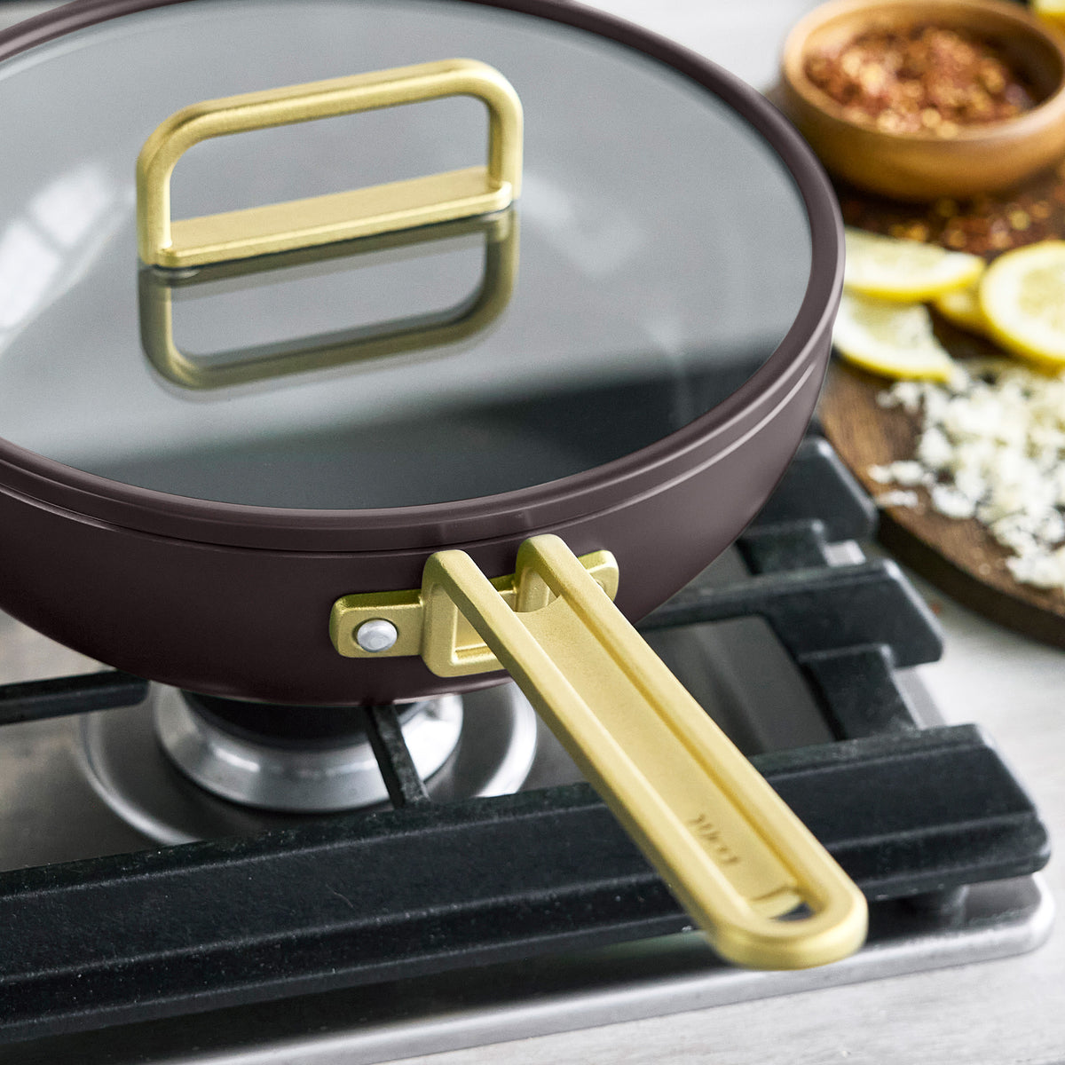 Espresso Stanley Tucci™ 3.25-quart ceramic nonstick piccolo pan with a glass lid and gold-toned handles on a stovetop.