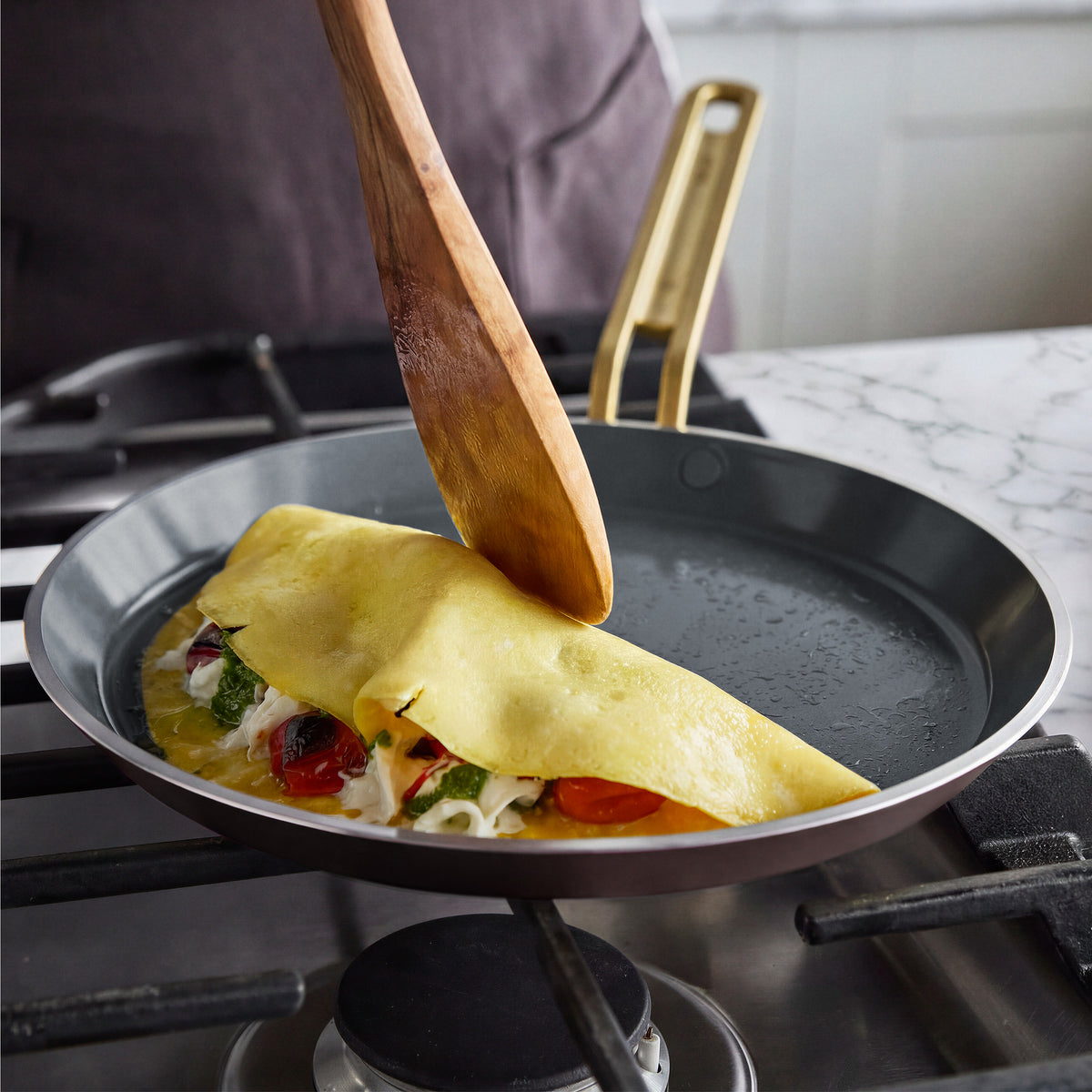 Wooden spatula folding an omelette in an espresso Stanley Tucci Ceramic Nonstick 10-inch crepe and omelette pan on a gas stovetop.