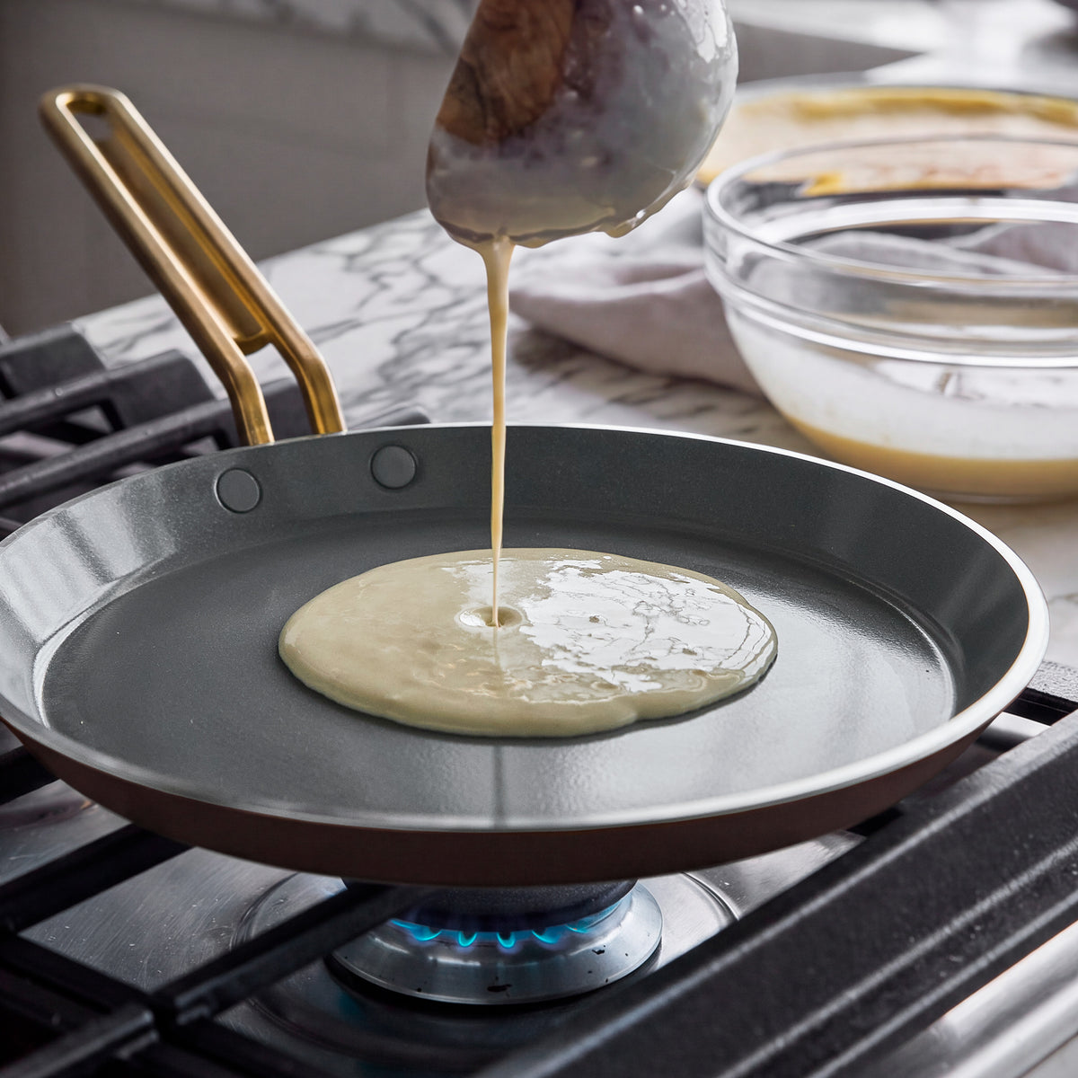 Batter pouring from a ladle into an espresso Stanley Tucci 10-inch ceramic nonstick crepe and omelette pan on a gas stove.