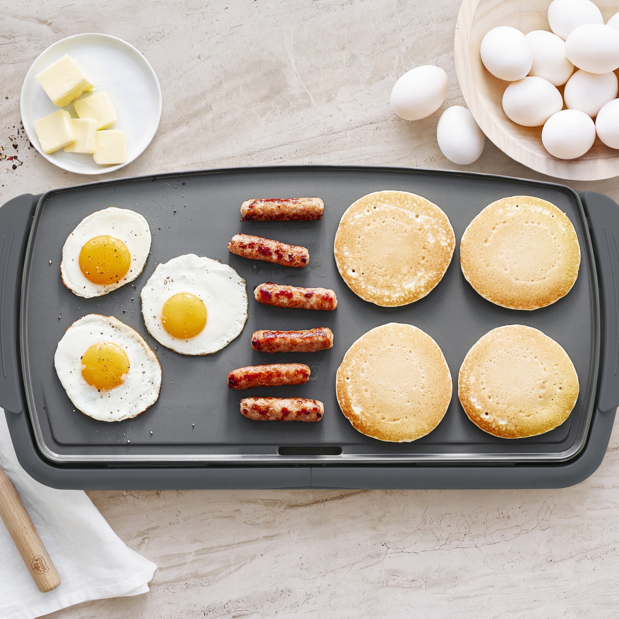 Pancakes, sausages, and eggs cooking on a graphite GreenLife Griddle XL with extra eggs and butter on the side.