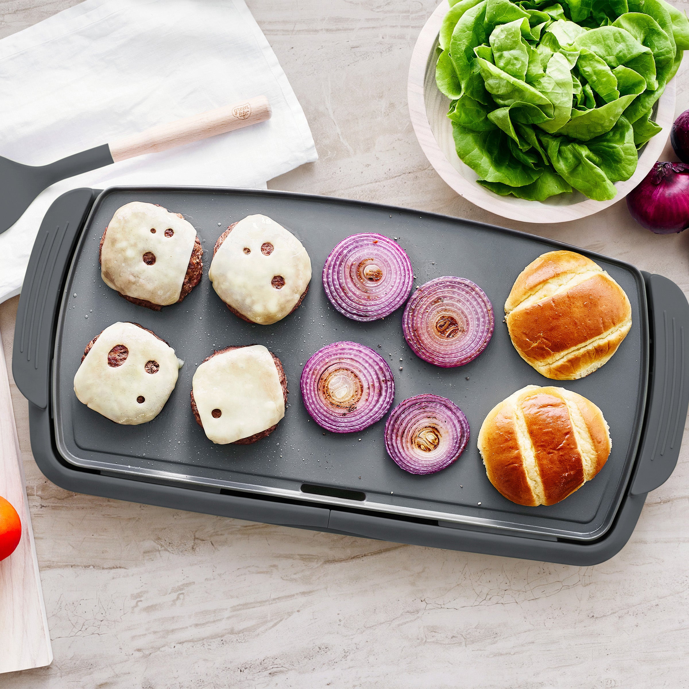 GreenLife XL graphite electric griddle with cheeseburgers, onions, buns, lettuce, and a spatula on a light countertop.