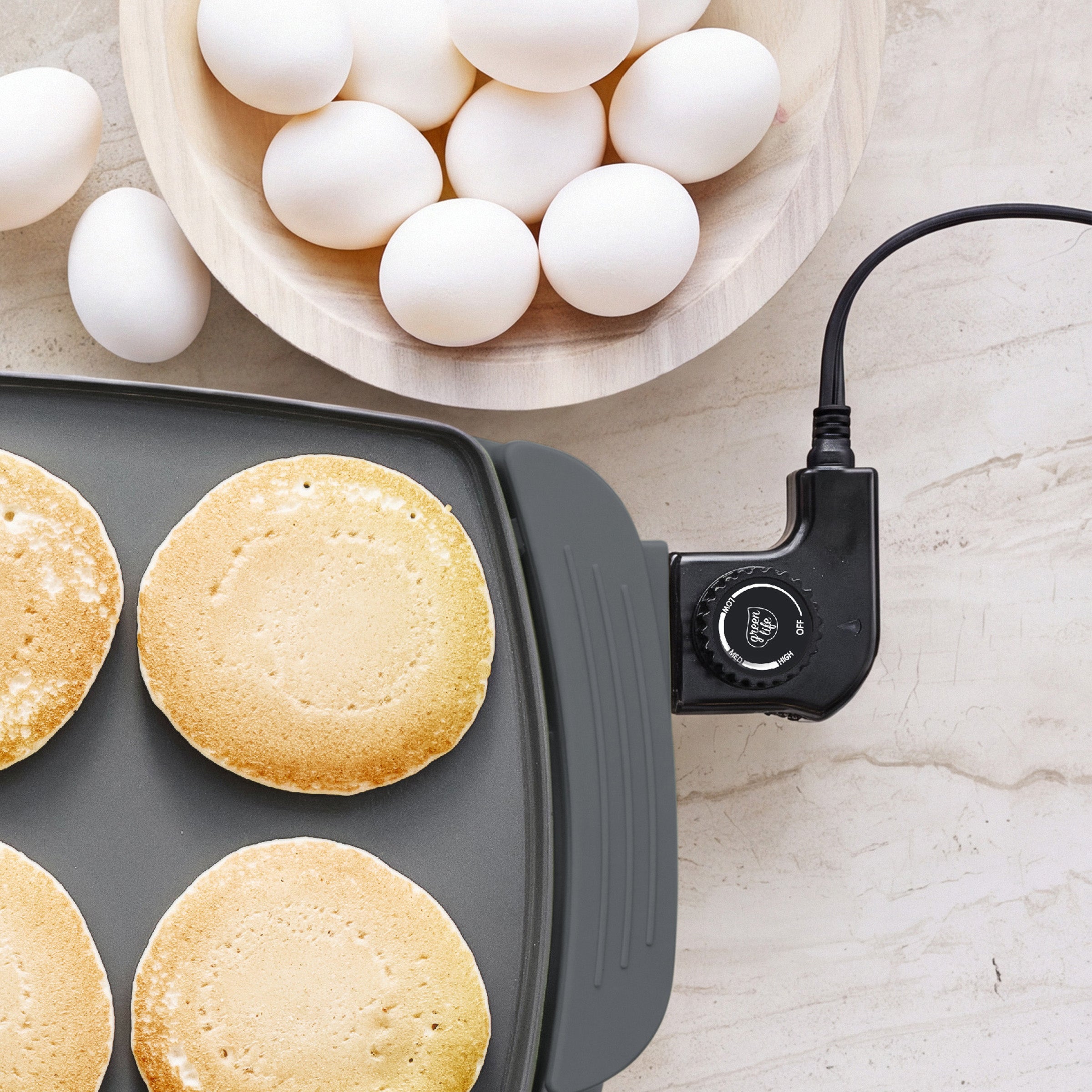 Four pancakes cooking on a graphite GreenLife Griddle XL with a bowl of eggs on a light countertop.