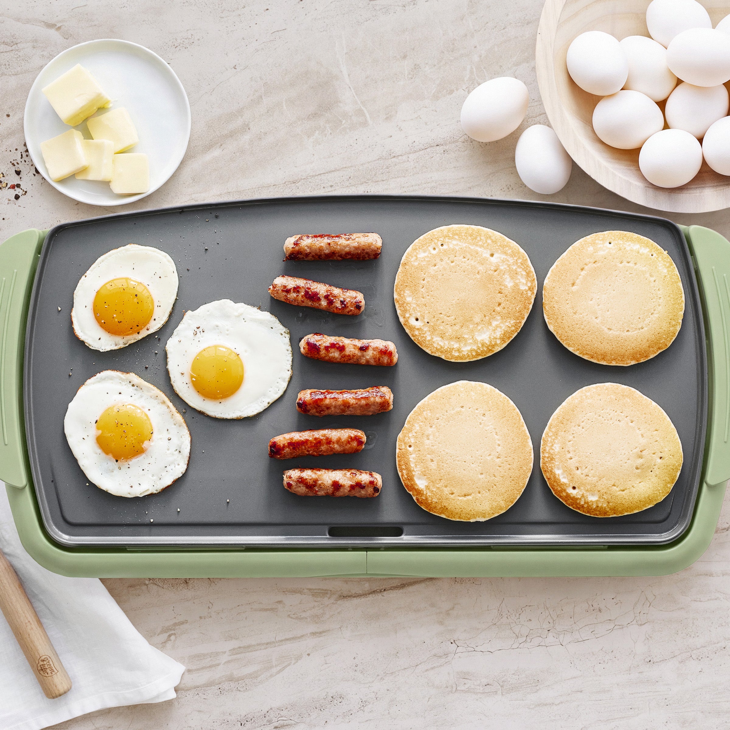 Sage green griddle with two fried eggs, five sausage links, and four pancakes on the cooking surface.