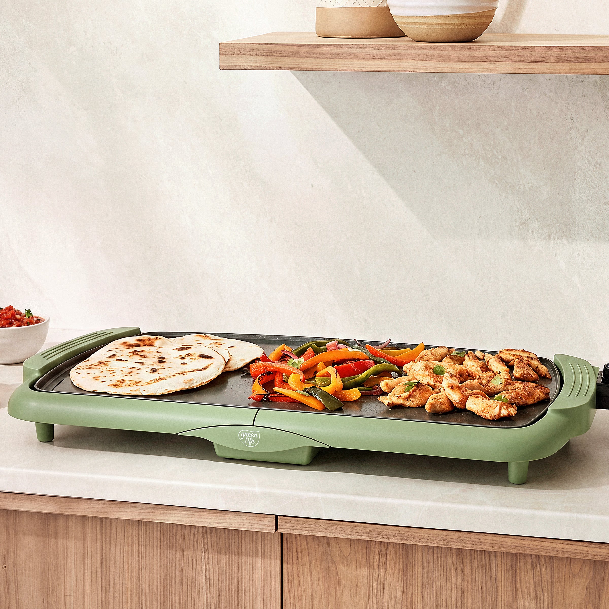 GreenLife Griddle XL on a kitchen counter with flatbread, sliced peppers, and chicken cooking beneath a wooden shelf.