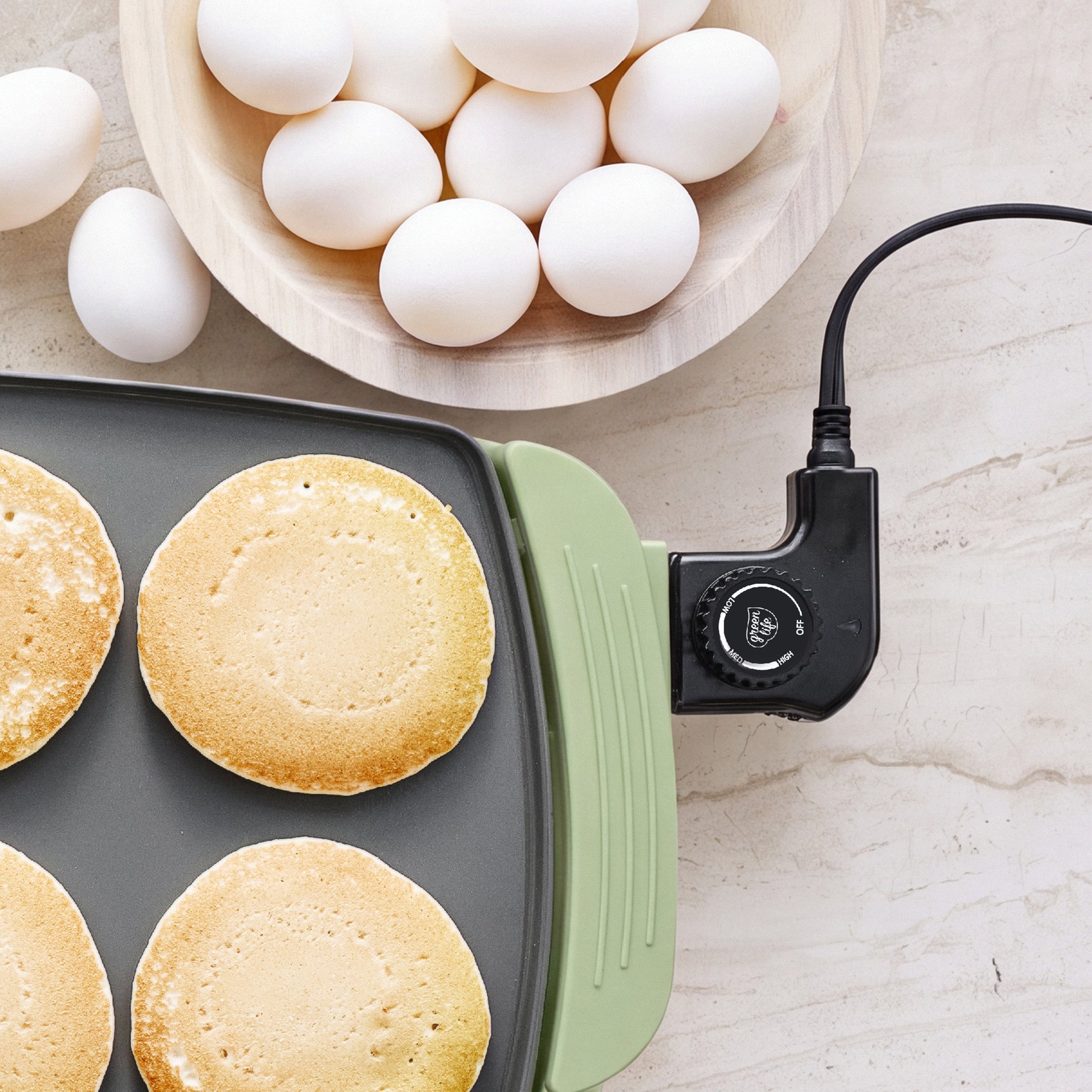 Four pancakes on a GreenLife Griddle XL with sage and a bowl of eggs on a light countertop.