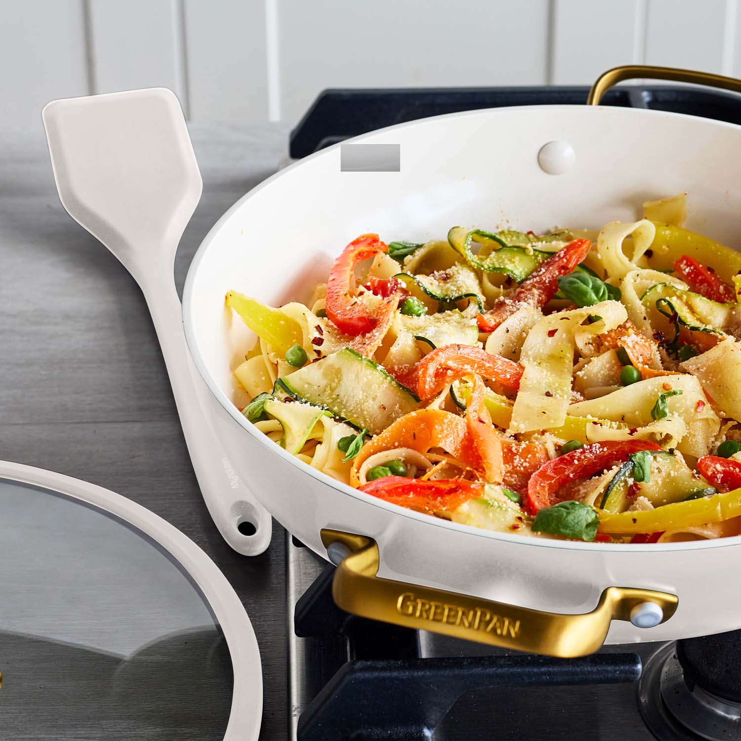 Cream ceramic pan with veggie pasta, basil, cheese, and a spatula on the stove.