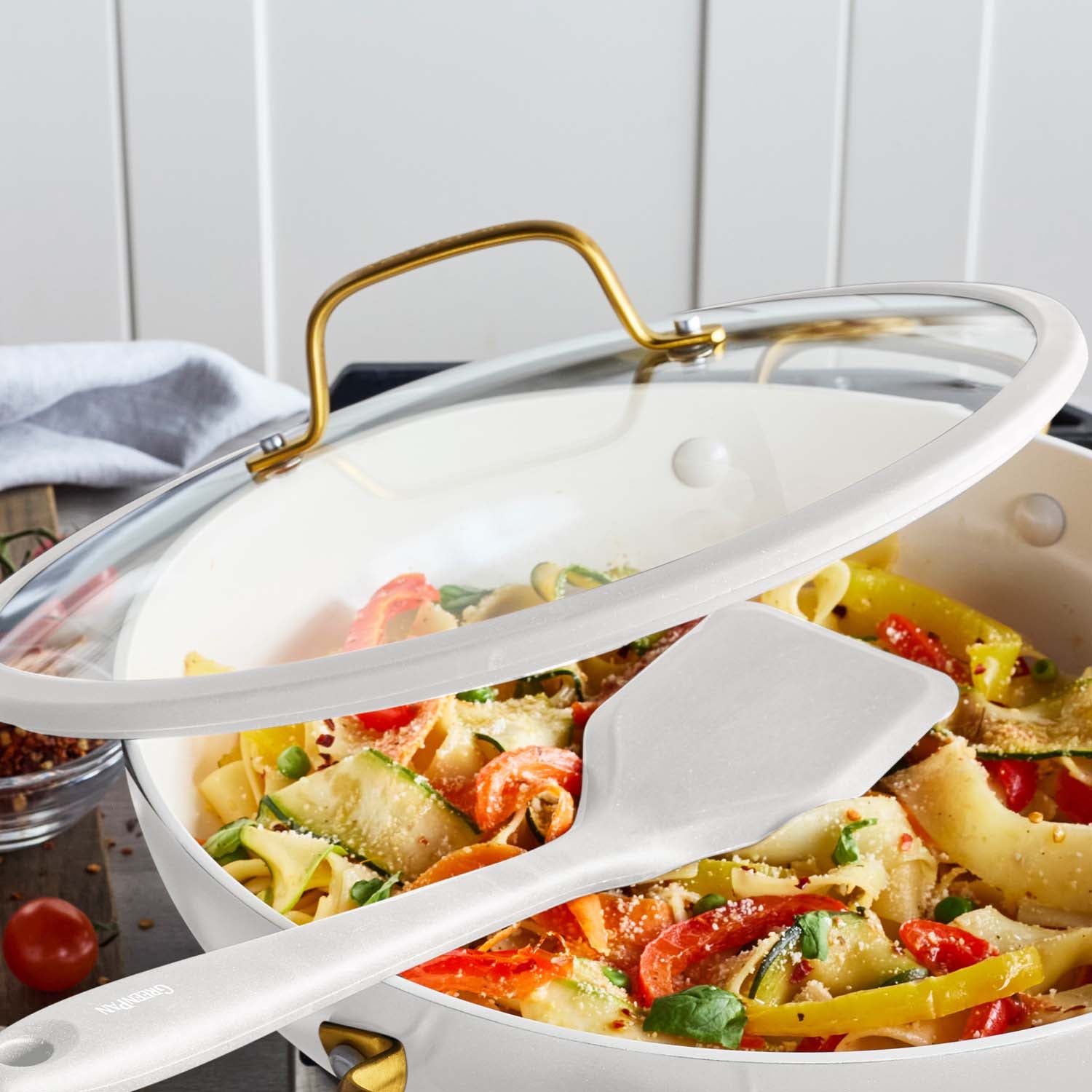 Cream ceramic nonstick everyday pan with lid, shown with cooked pasta inside; a white spatula rests beside the pan.