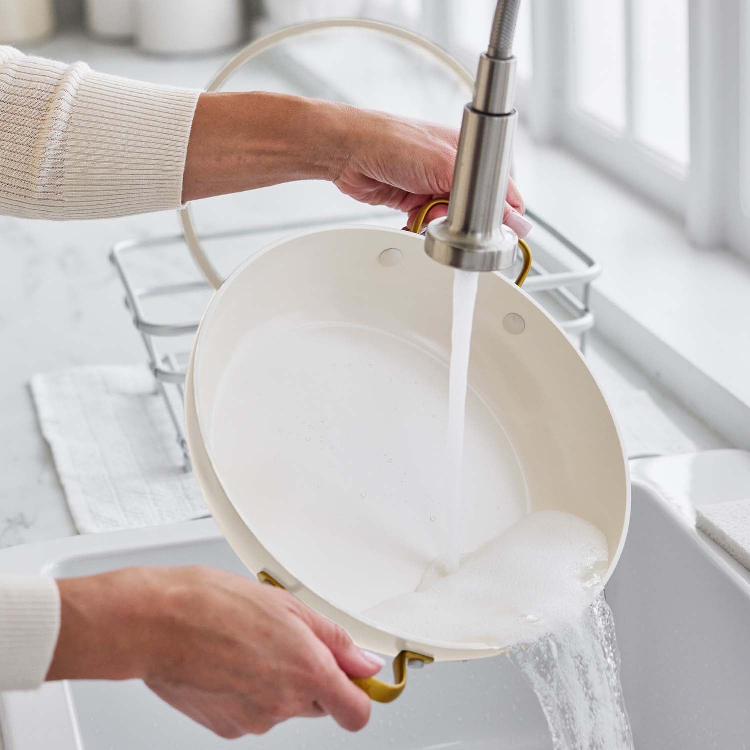 Cream ceramic nonstick pan with lid being rinsed under running water in a bright kitchen sink.