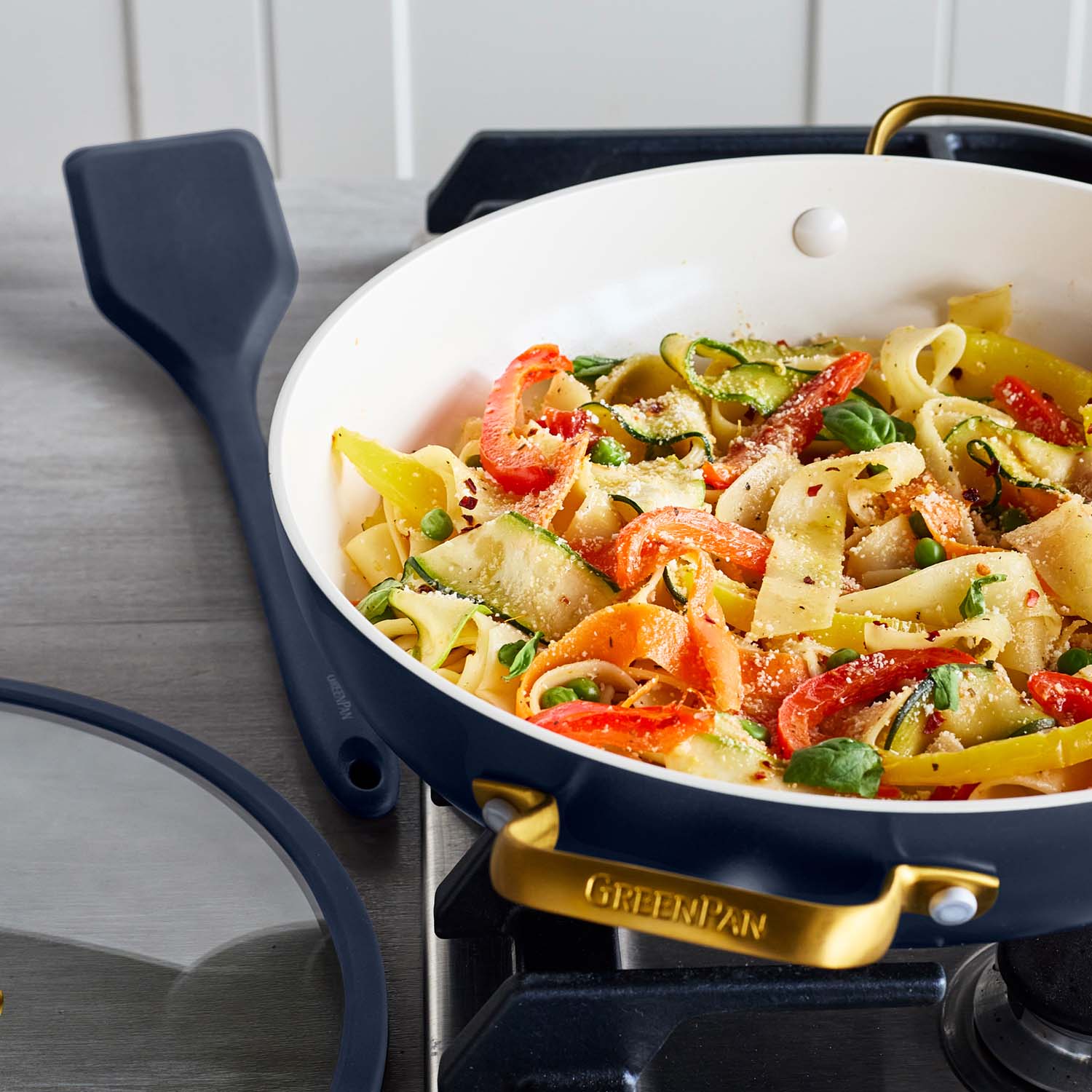 Blue ceramic nonstick everyday pan with veggie ribbon pasta and greens, spatula resting beside it on the stove.