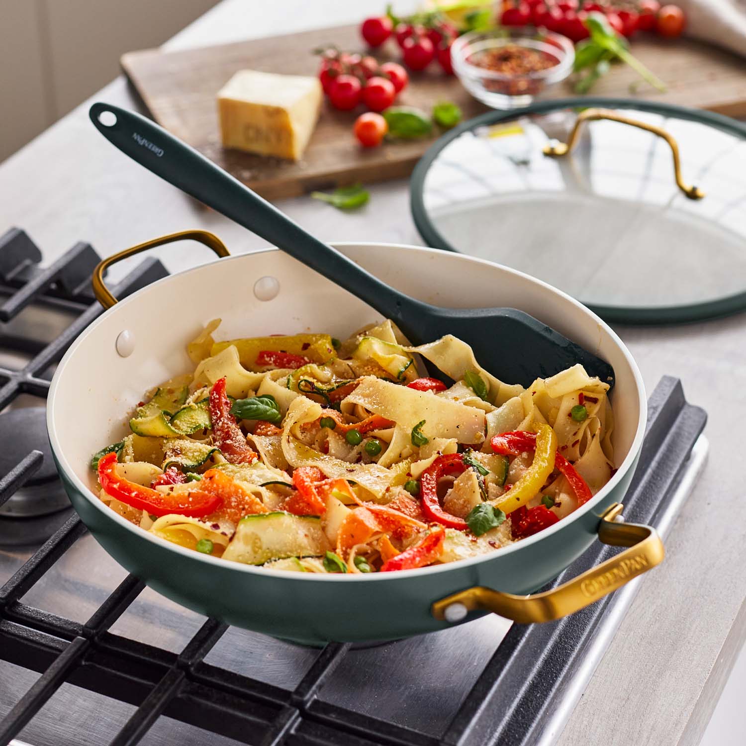 Vegetable pasta cooking in a green ceramic nonstick pan with a spatula and fresh veggies alongside.