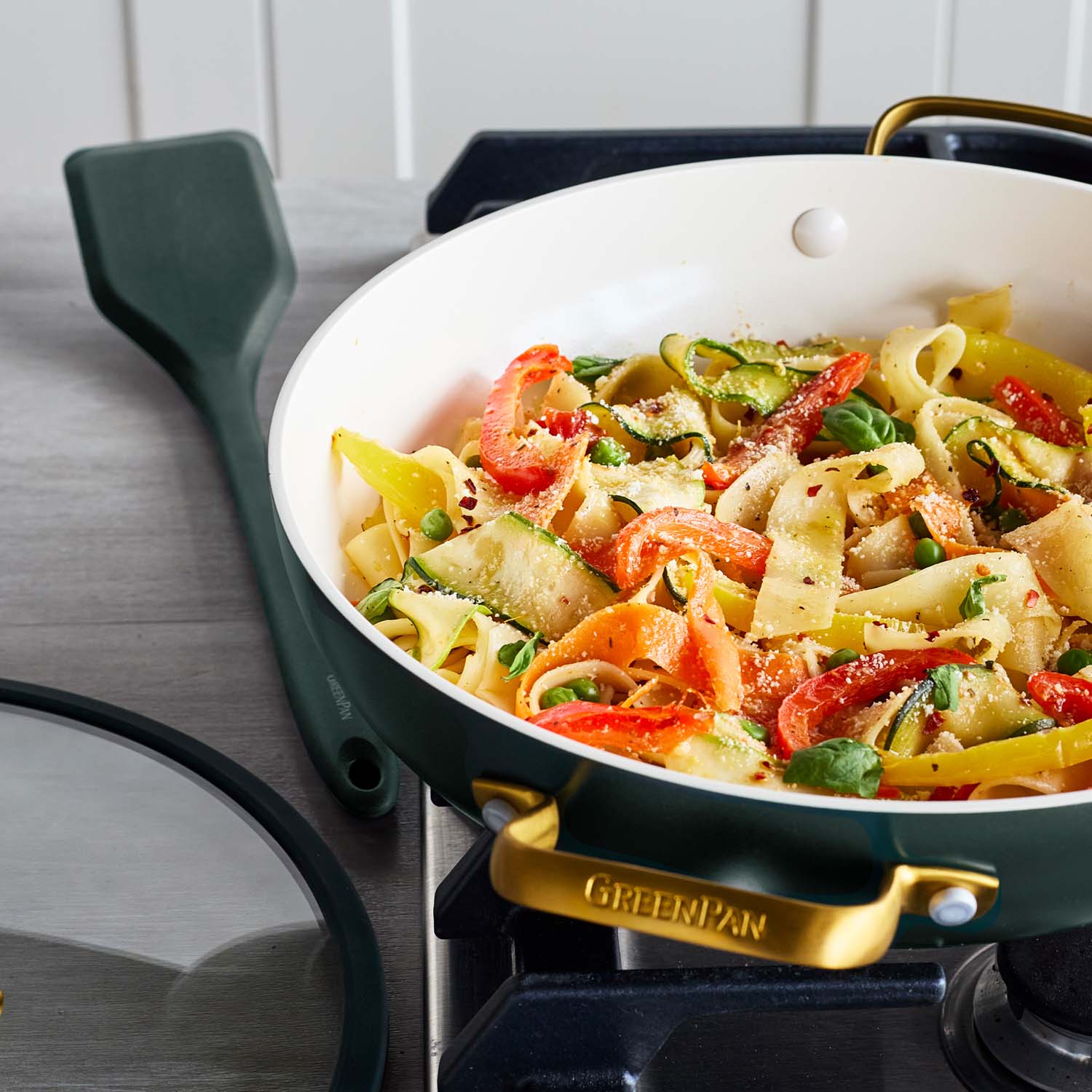 Vegetable ribbon pasta in a green ceramic nonstick pan with lid, shown on a stovetop and garnished.