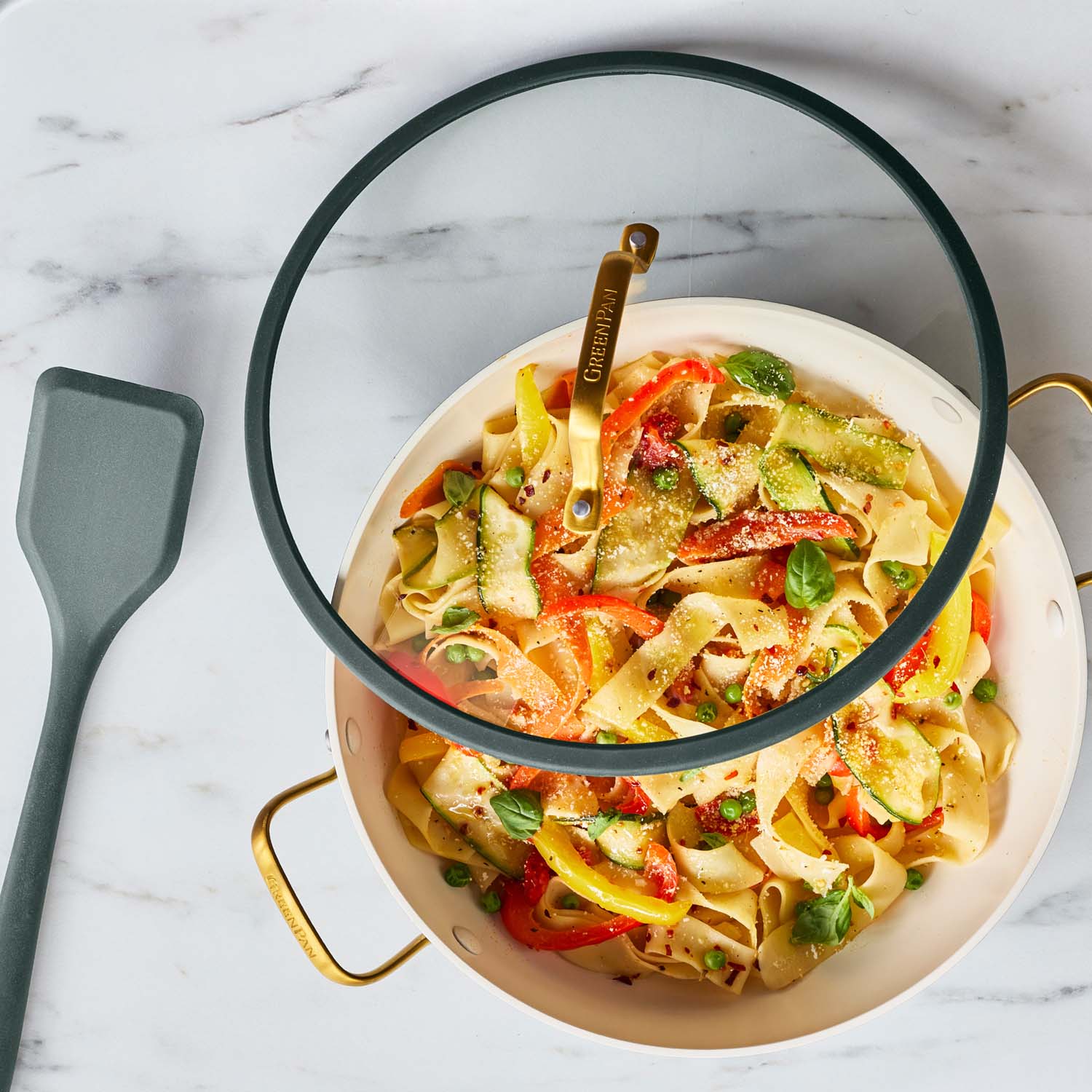 Green ceramic nonstick 3.75-qt pan with lid, filled with veggie pasta, beside a gray spatula on a marble surface.