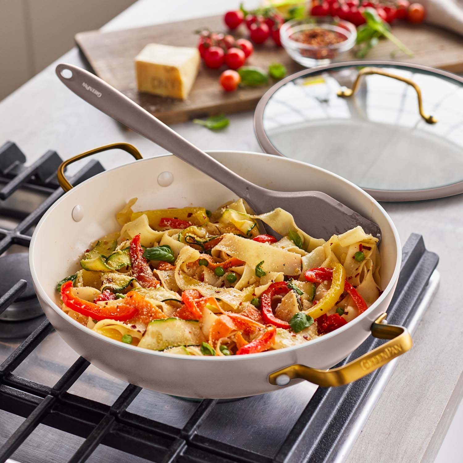 Cream-colored ceramic nonstick pan with lid on stove, cooking pasta and vegetables; spatula and ingredients beside pan.