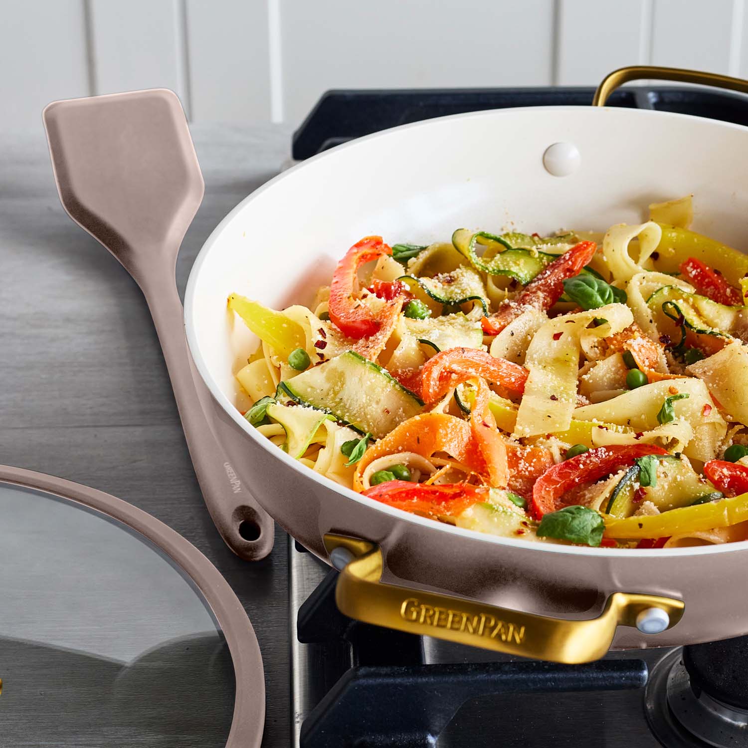 Veggie pasta cooking in a beige ceramic nonstick pan with glass lid, spatula resting beside on the countertop.