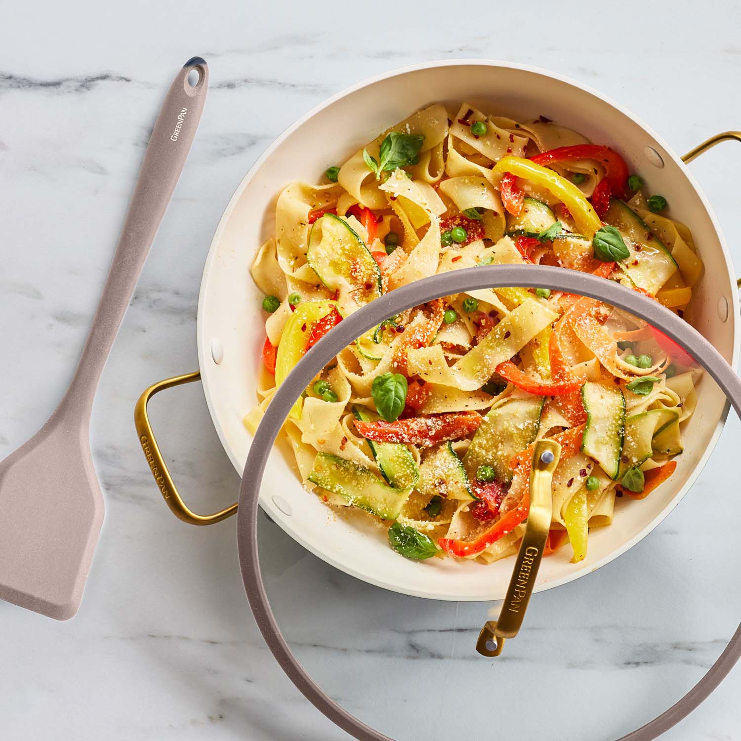 Veggie pasta in a beige ceramic nonstick pan with lid, alongside a beige spatula on marble surface.