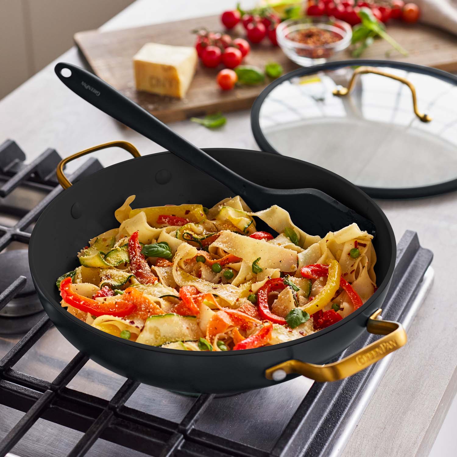 Black ceramic everyday pan with lid and spatula, filled with vegetable pasta, fresh ingredients arranged nearby.