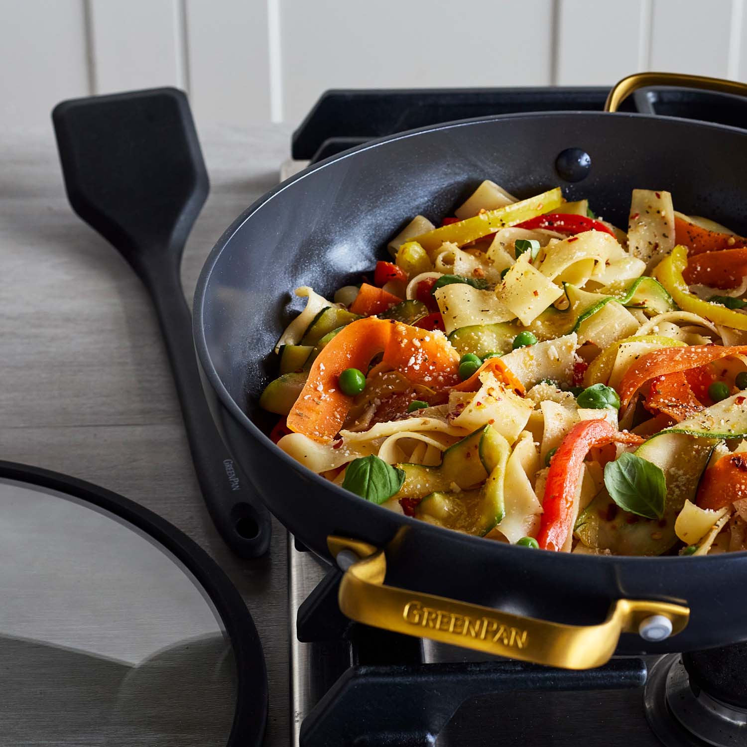 Black ceramic nonstick everyday pan with lid on stove, filled with pasta, peas, and mixed vegetables.