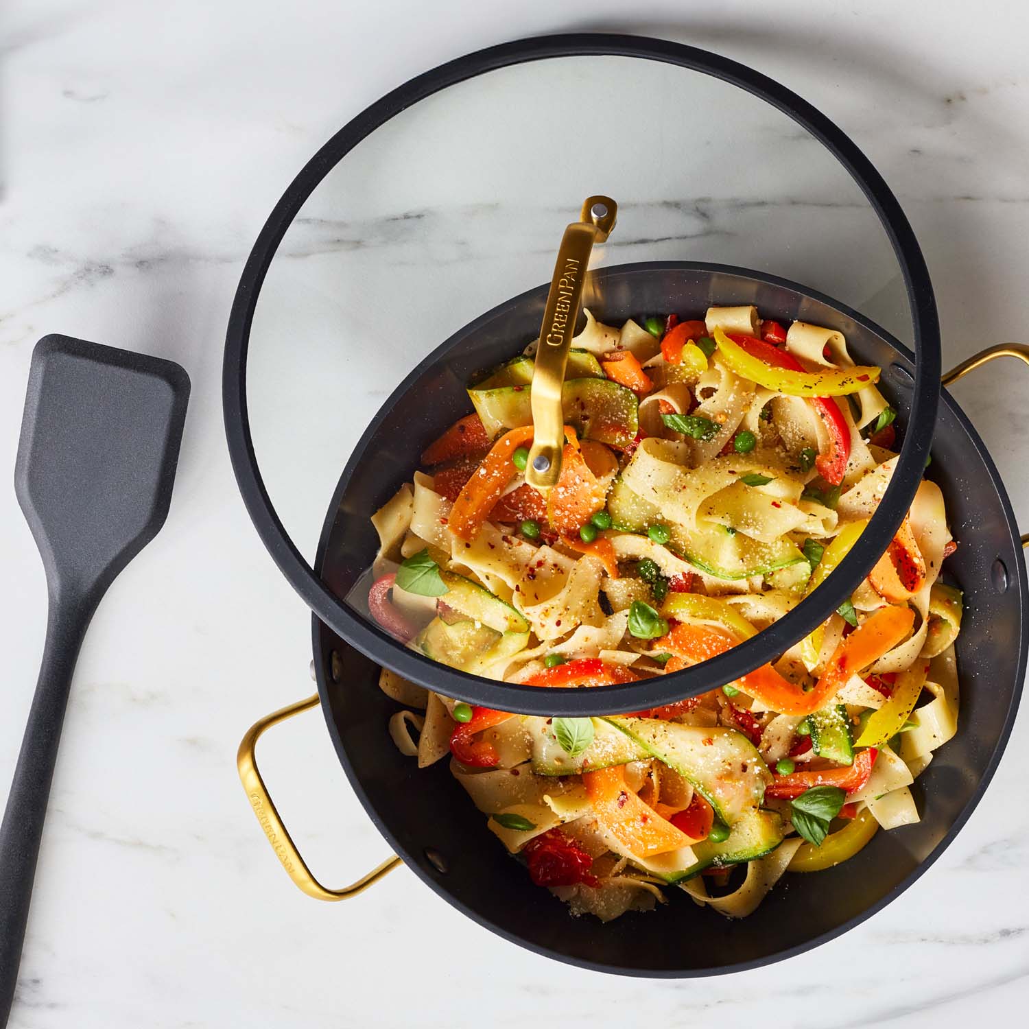 Black ceramic everyday pan with lid, open and filled with veggie pasta and a spatula, on a white marble countertop.