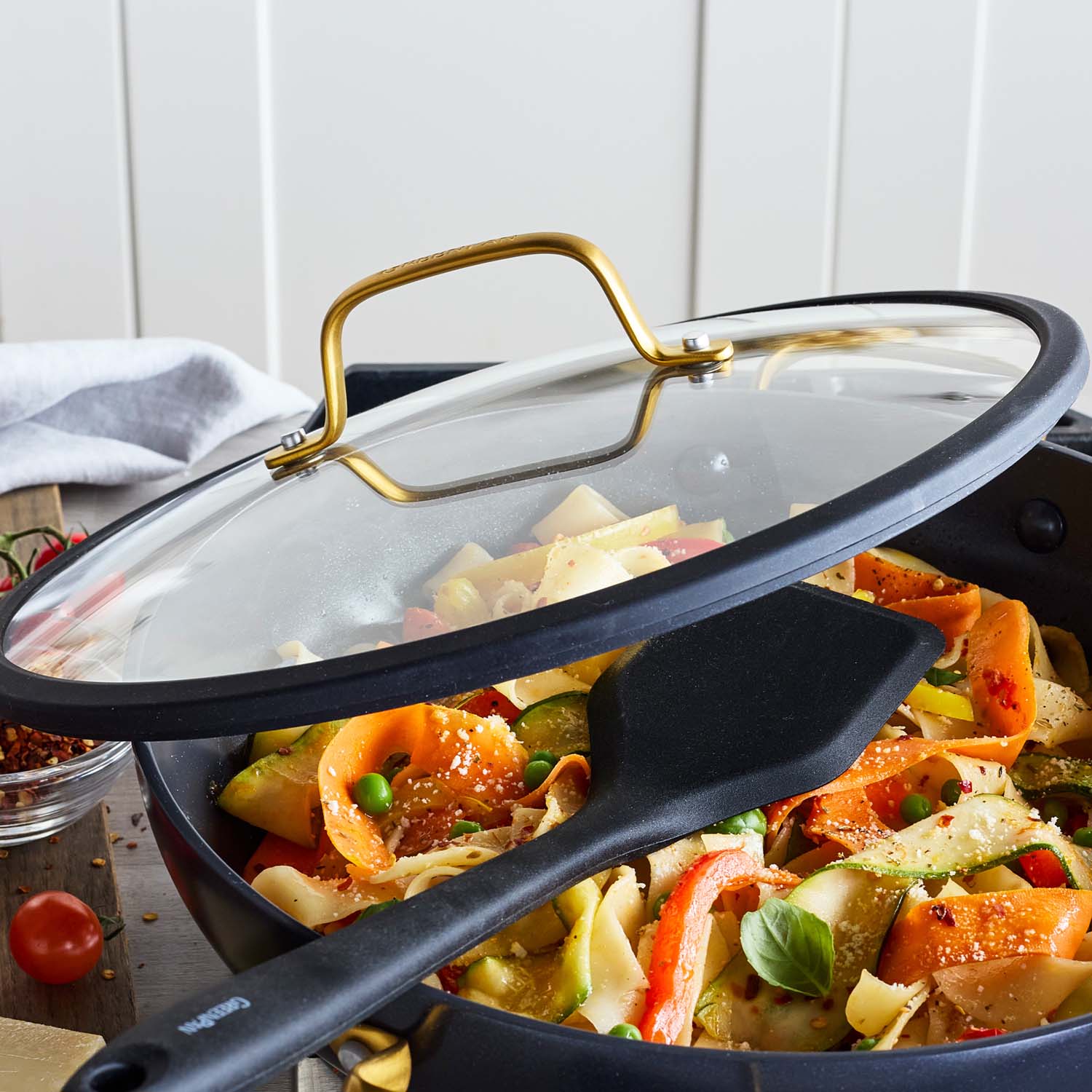 Black ceramic nonstick everyday pan with lid holding veggie pasta and a spatula, set on a table near tomatoes and a napkin.