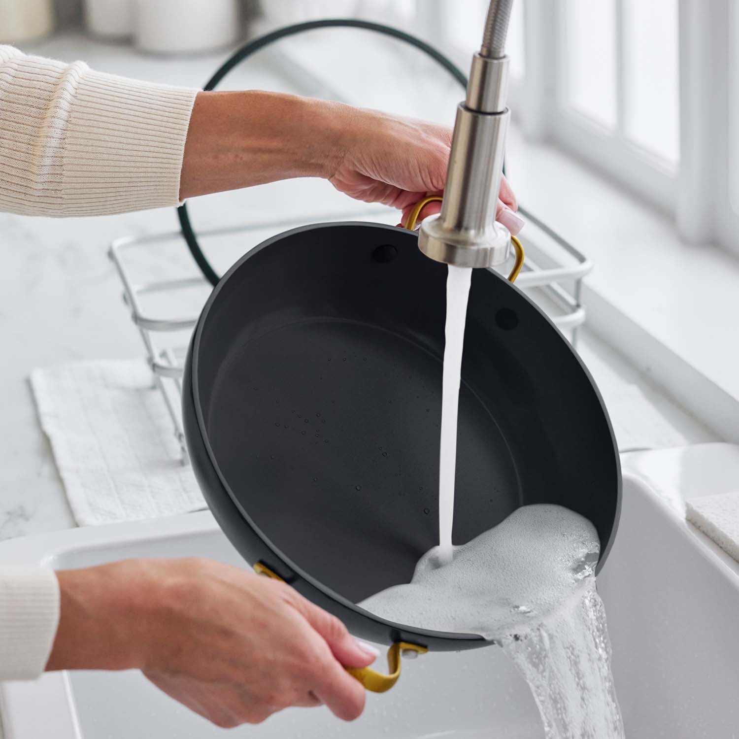 Washing a ceramic nonstick 3.75-quart pan with lid under running water in a sink, soap suds rinsing off.