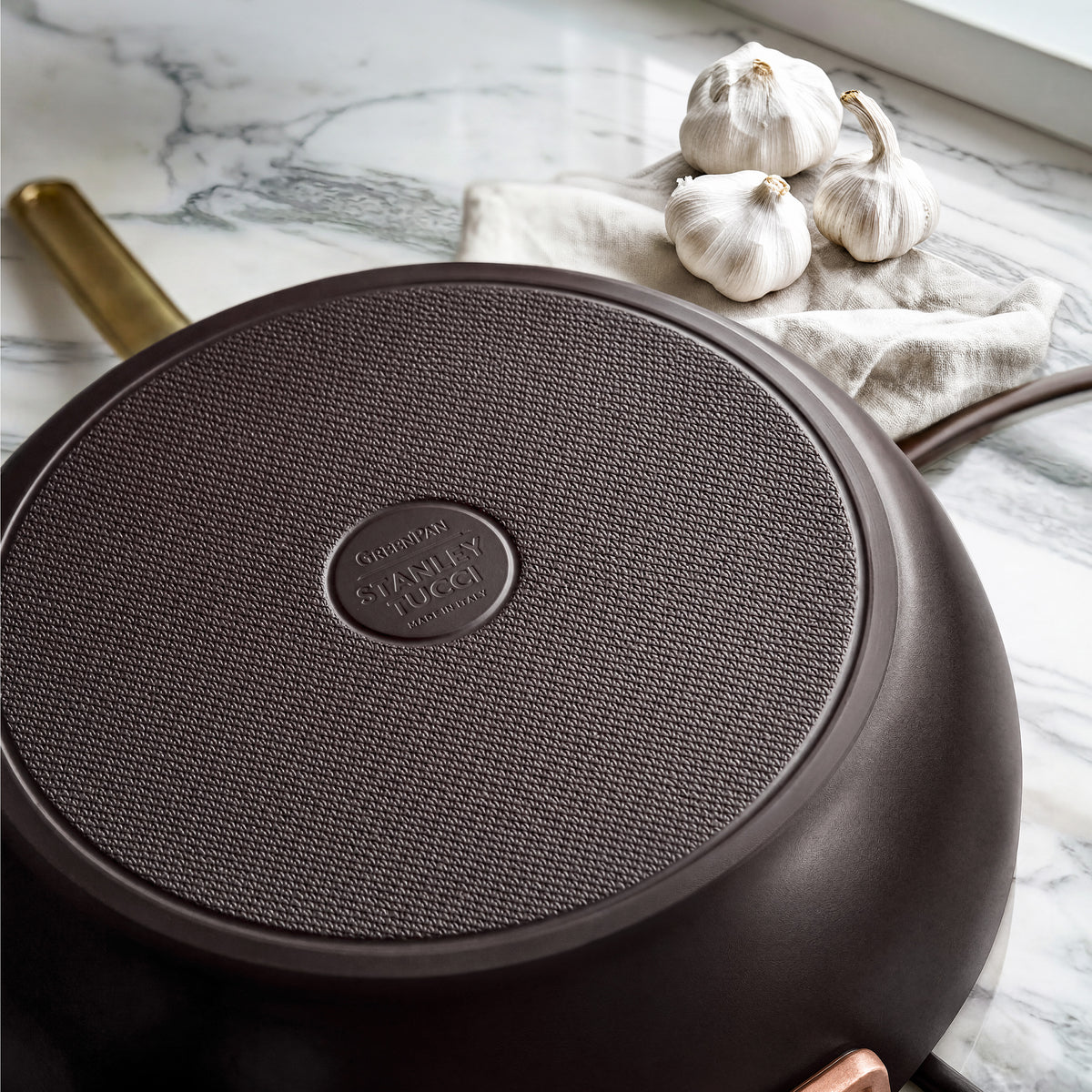 Espresso Stanley Tucci ceramic nonstick 6.5-quart essential pan with a textured base on a marble surface next to three garlic bulbs.