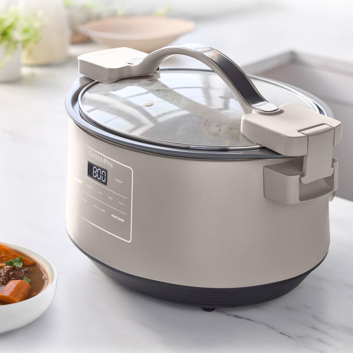 Clay-colored slow cooker with locking lid on a kitchen counter, next to a bowl of stew.