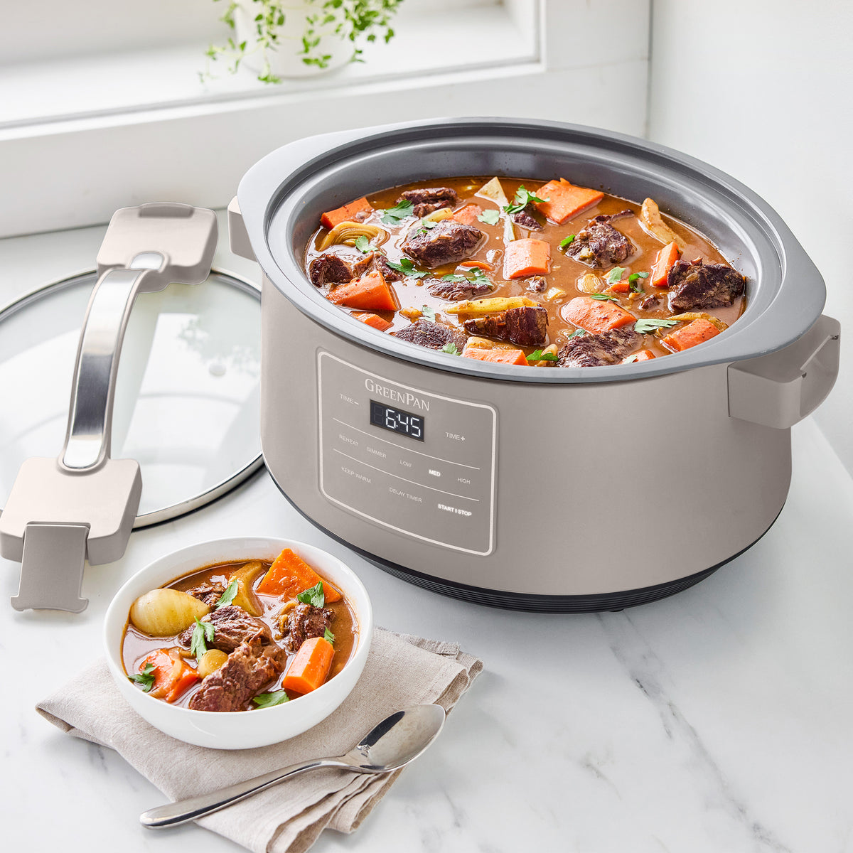 Clay slow cooker with locking lid on a white counter, filled with beef stew. Lid and bowl of stew are beside the cooker.