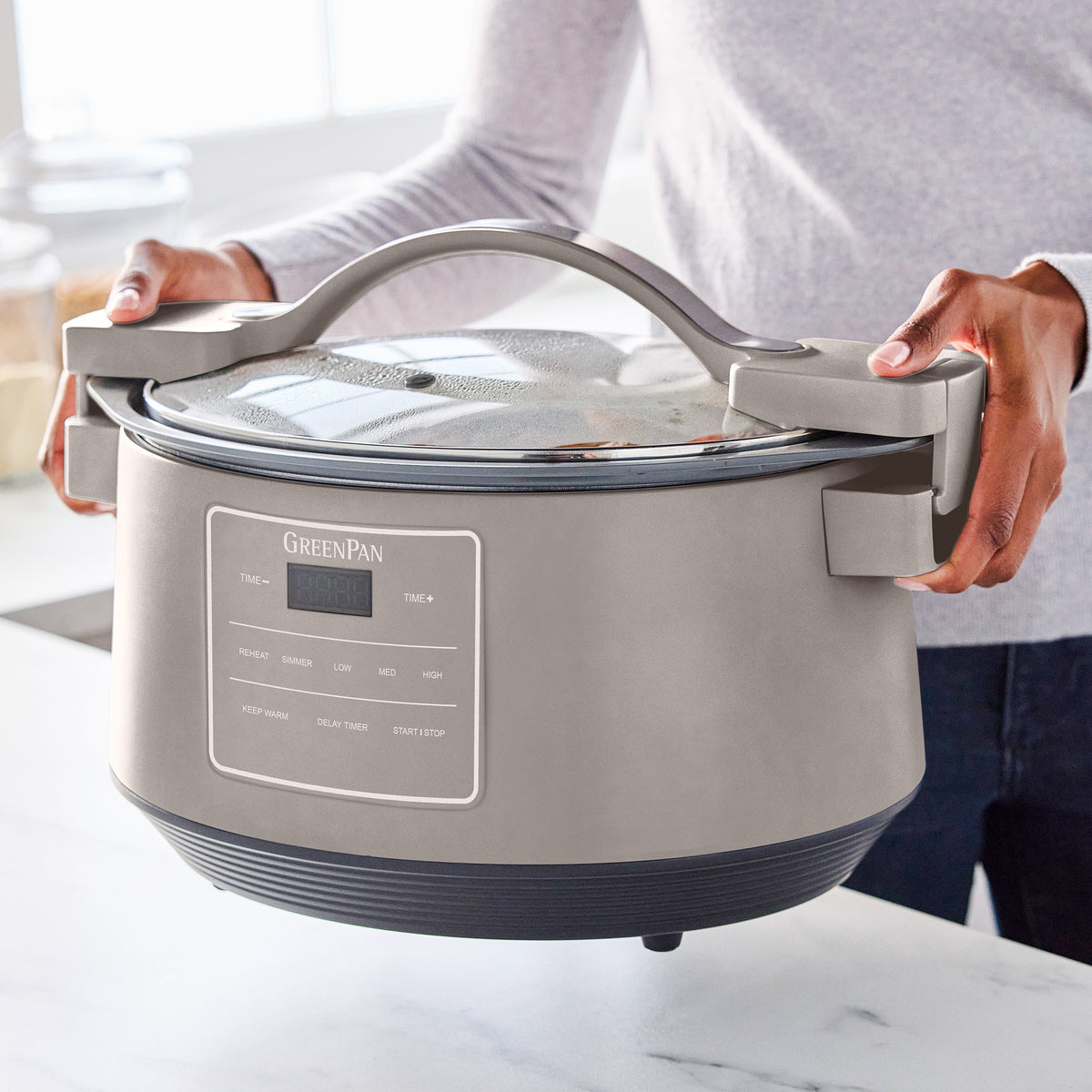 Hands holding an 8-quart clay slow cooker with a locking glass lid in a bright kitchen.