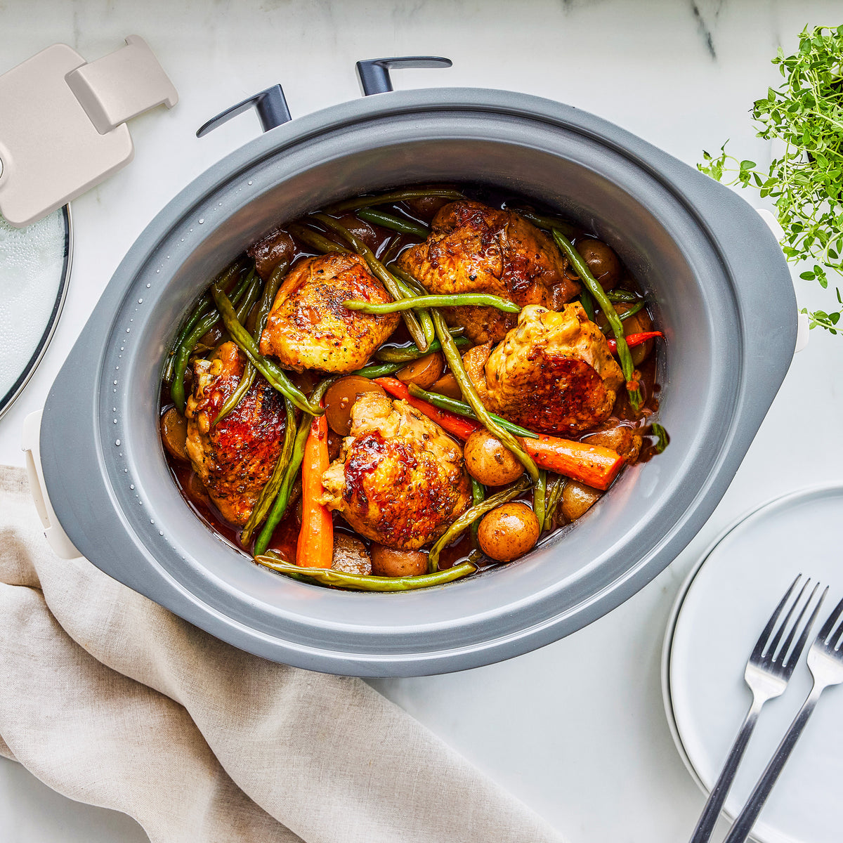 Slow cooker with locking lid, clay pot filled with chicken, green beans, carrots, and potatoes on a white table.