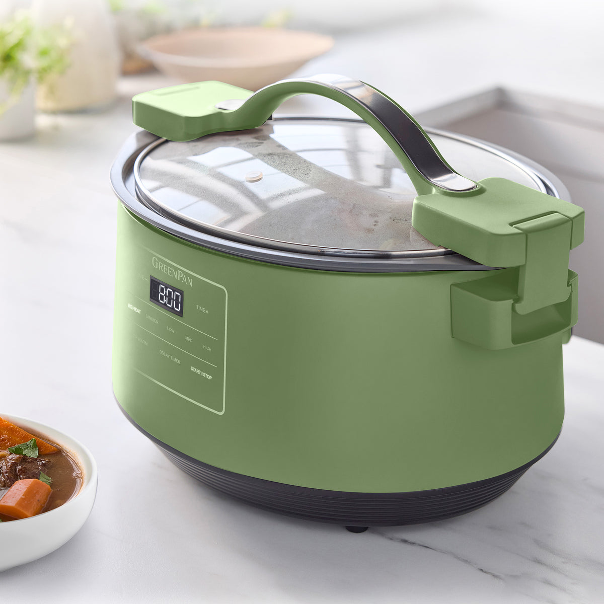 Sage green 8-quart slow cooker with locking lid on a counter next to a bowl of stew.