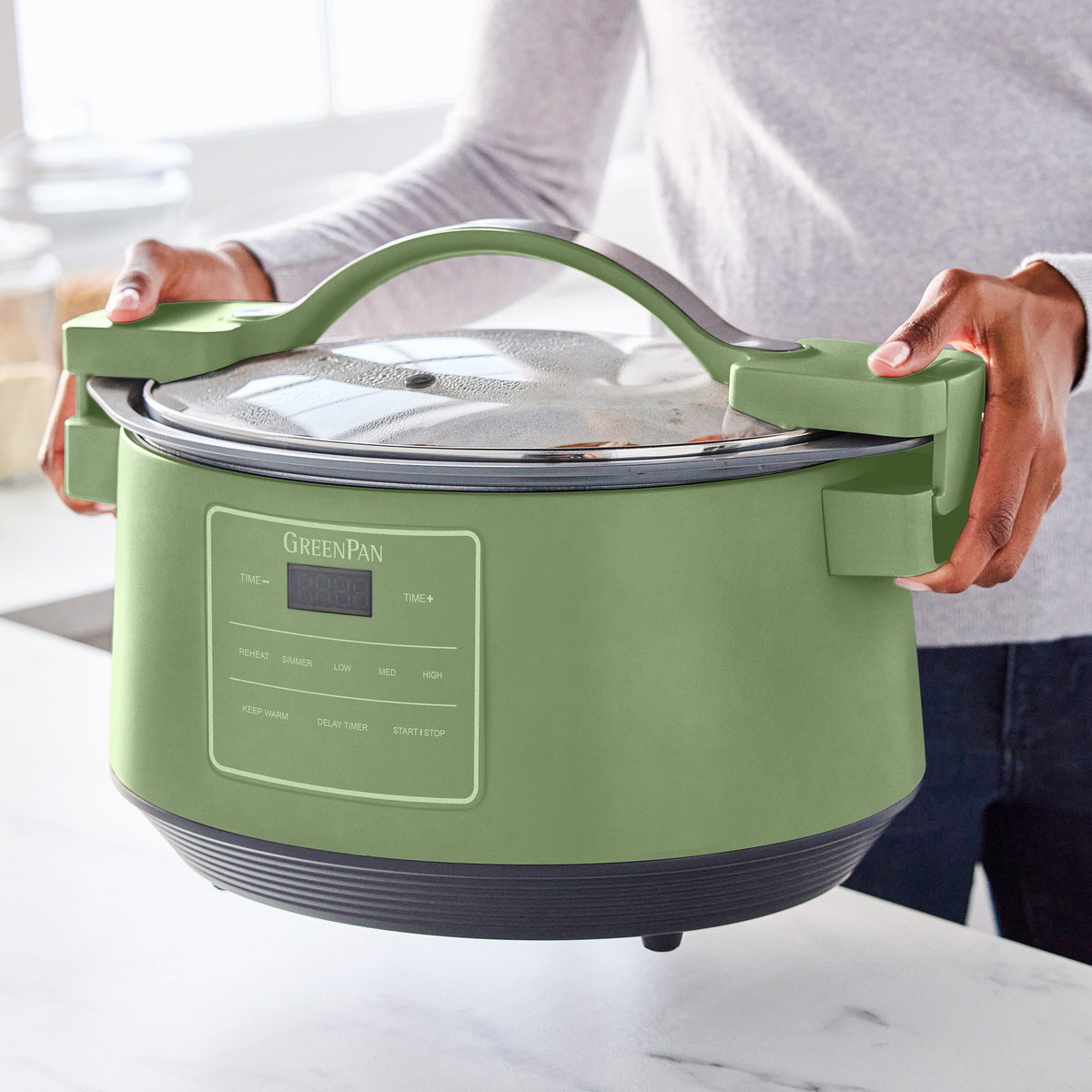 Person holding sage green 8-quart slow cooker with clear locking lid in a well-lit kitchen.