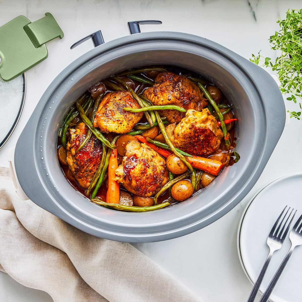 Sage green slow cooker with locking lid, filled with roasted chicken, potatoes, green beans, and carrots on a white surface.