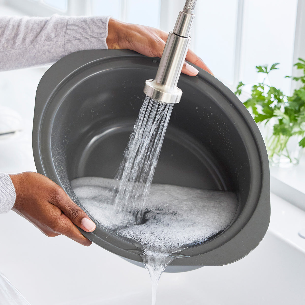 Person washing a sage green 8-quart slow cooker under a kitchen faucet; leafy plants sit by the window in the background.