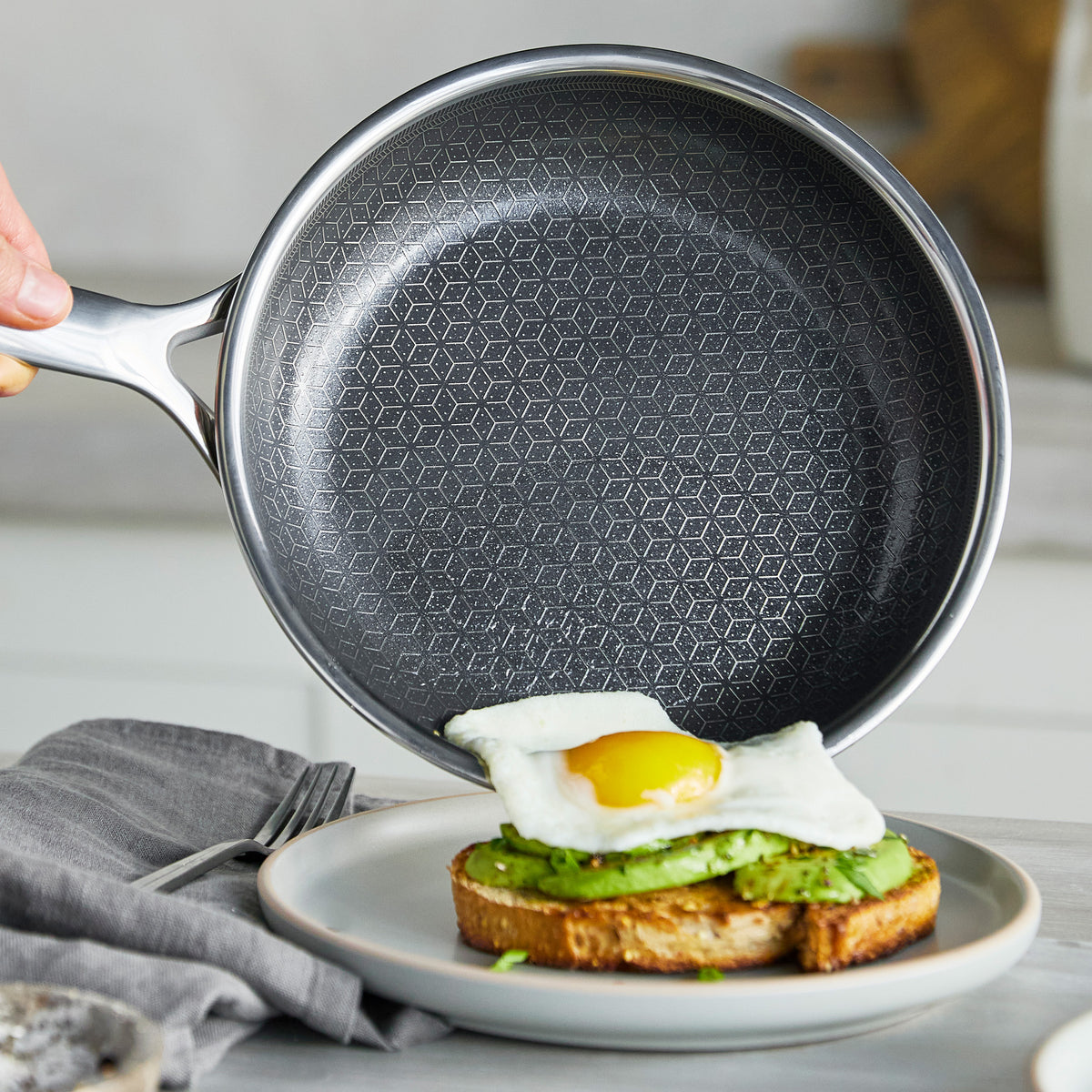 Hand tipping a gray ceramic 10-inch frypan to slide a sunny-side-up egg onto avocado toast.