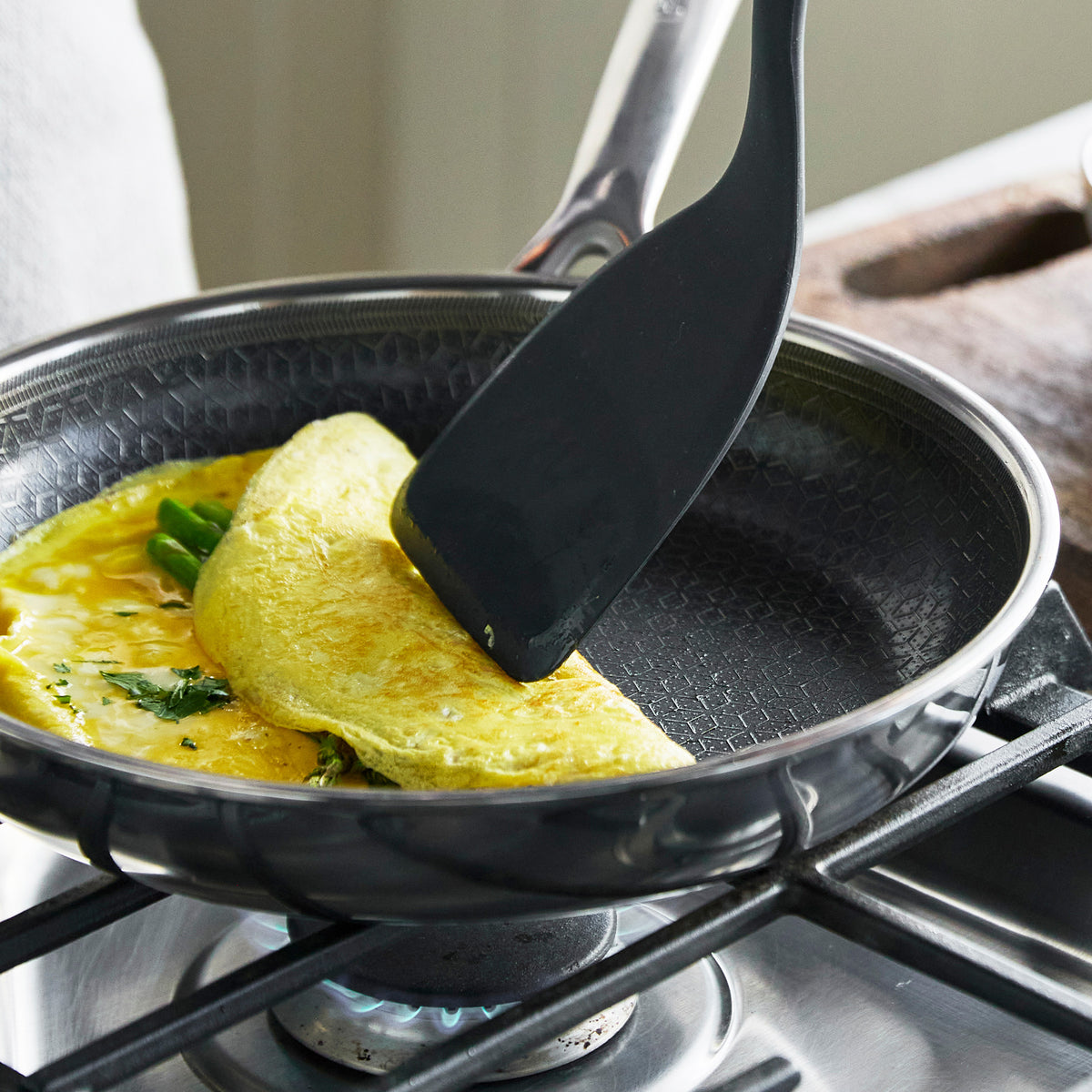 A black spatula folding an herbed omelet in a white ceramic nonstick frypan on a stovetop.