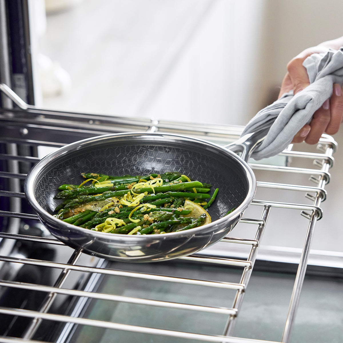 Hand with a towel holds a ceramic nonstick frypan filled with sautéed vegetables on an oven rack.