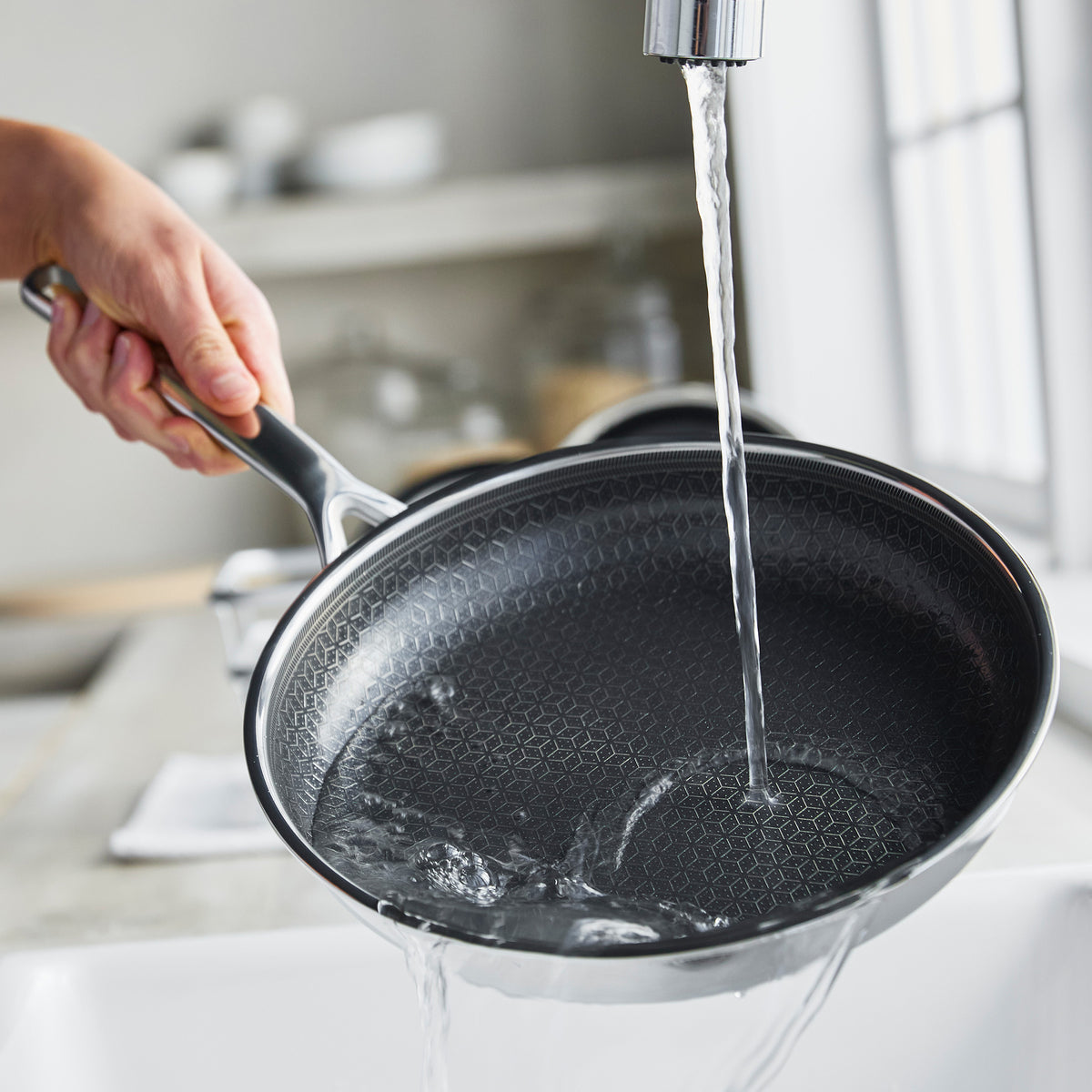 Person washing a gray ceramic nonstick 10-inch frypan by hand under running water in a kitchen sink.