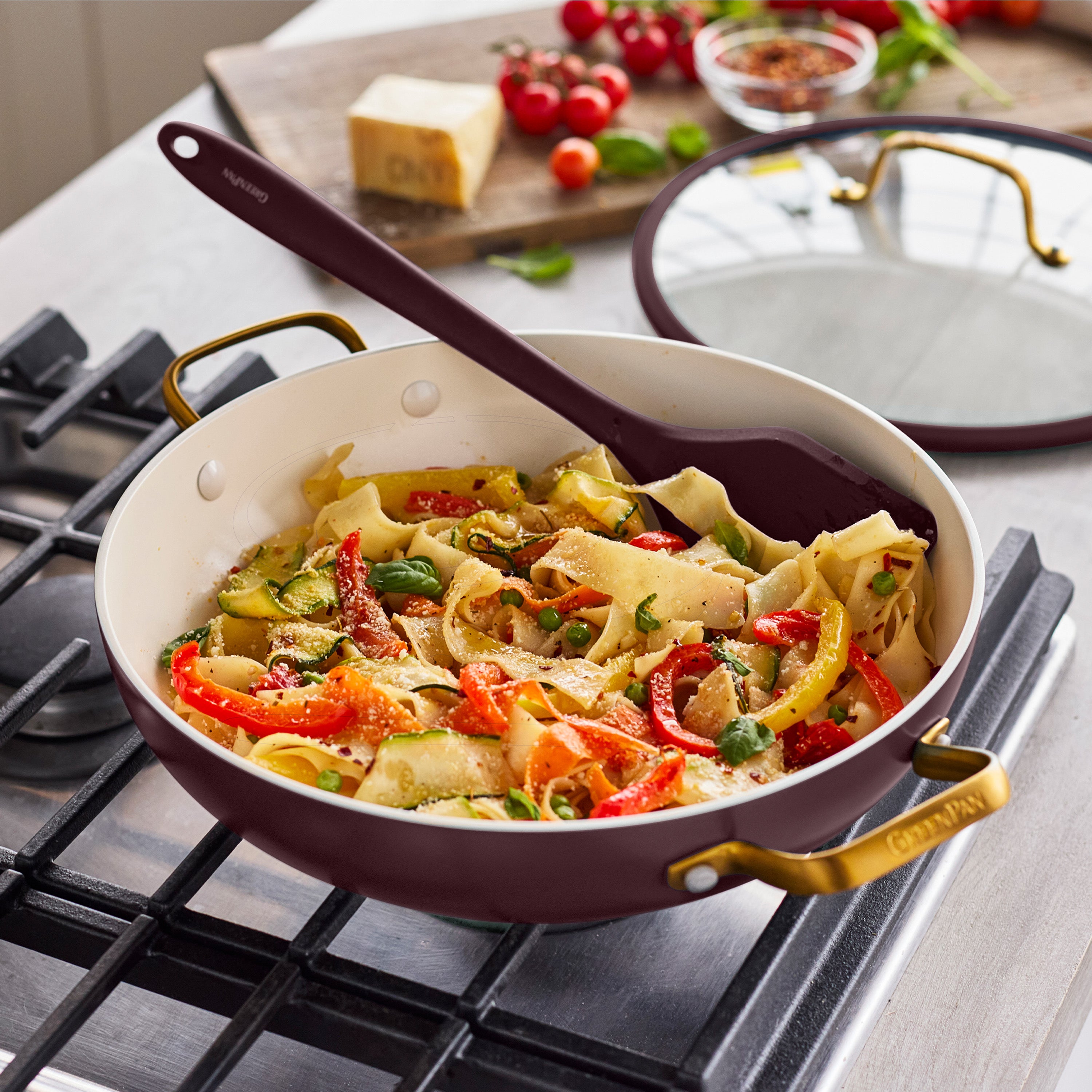 Bordeaux ceramic nonstick everyday pan with lid next to a skillet of pasta and veggies on the stove with a spatula.