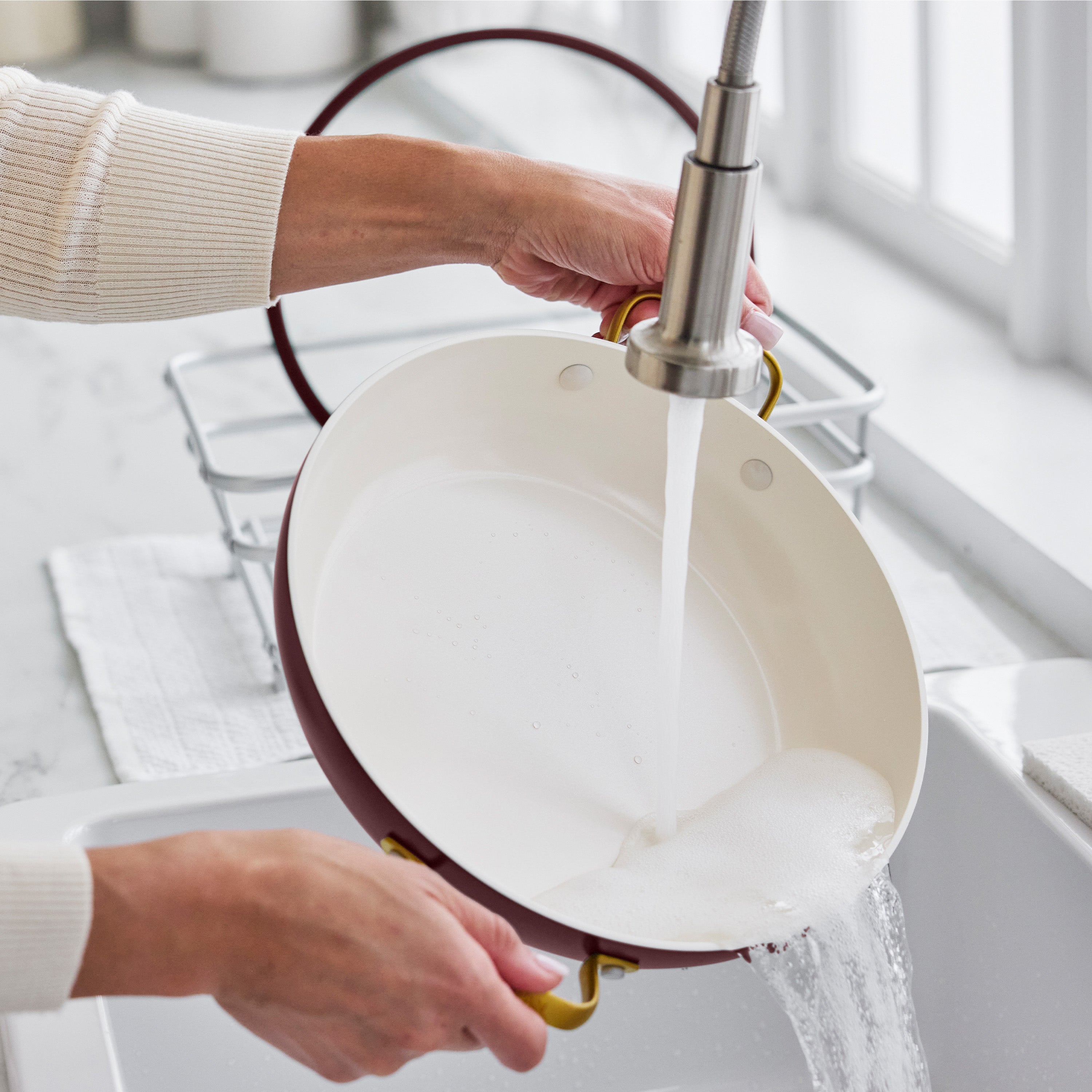 A soapy Bordeaux ceramic nonstick pan with lid is being rinsed at a sunny kitchen sink.