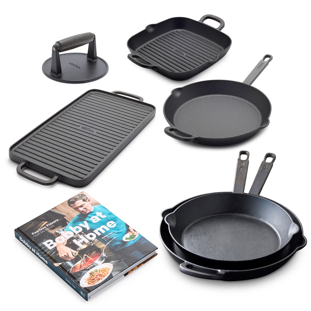 The Bobby Flay by GreenPan 6-Piece Cast Iron Set includes heirloom-quality black pans—a grill press, grill pan, griddle, two frying pans—and the "Bobby at Home" cookbook featuring a man on the cover.